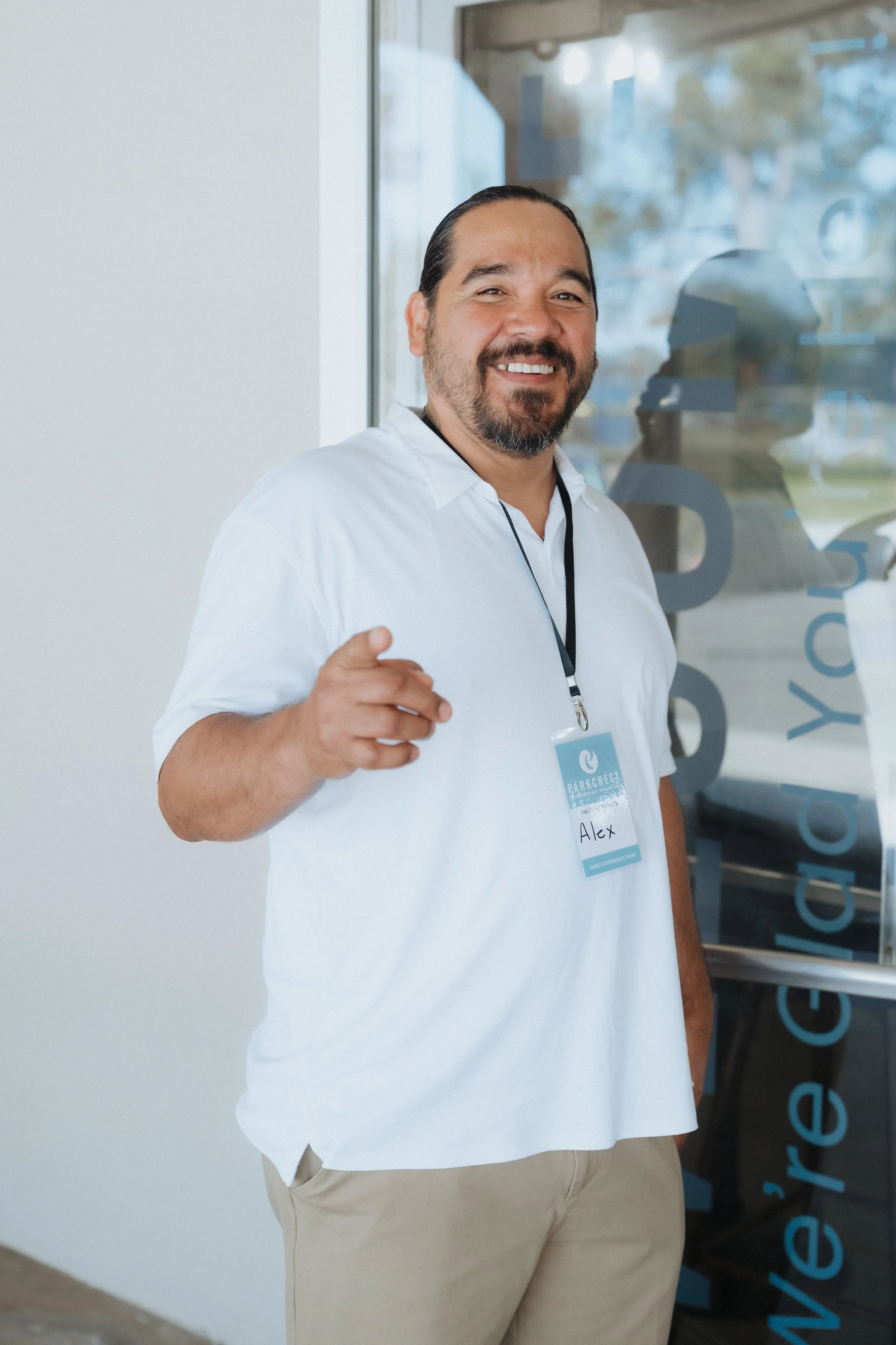 A smiling man wearing a white shirt and beige pants, standing outside a building with a glass door, pointing towards the camera, with a name badge that reads 'Alex' around his neck.