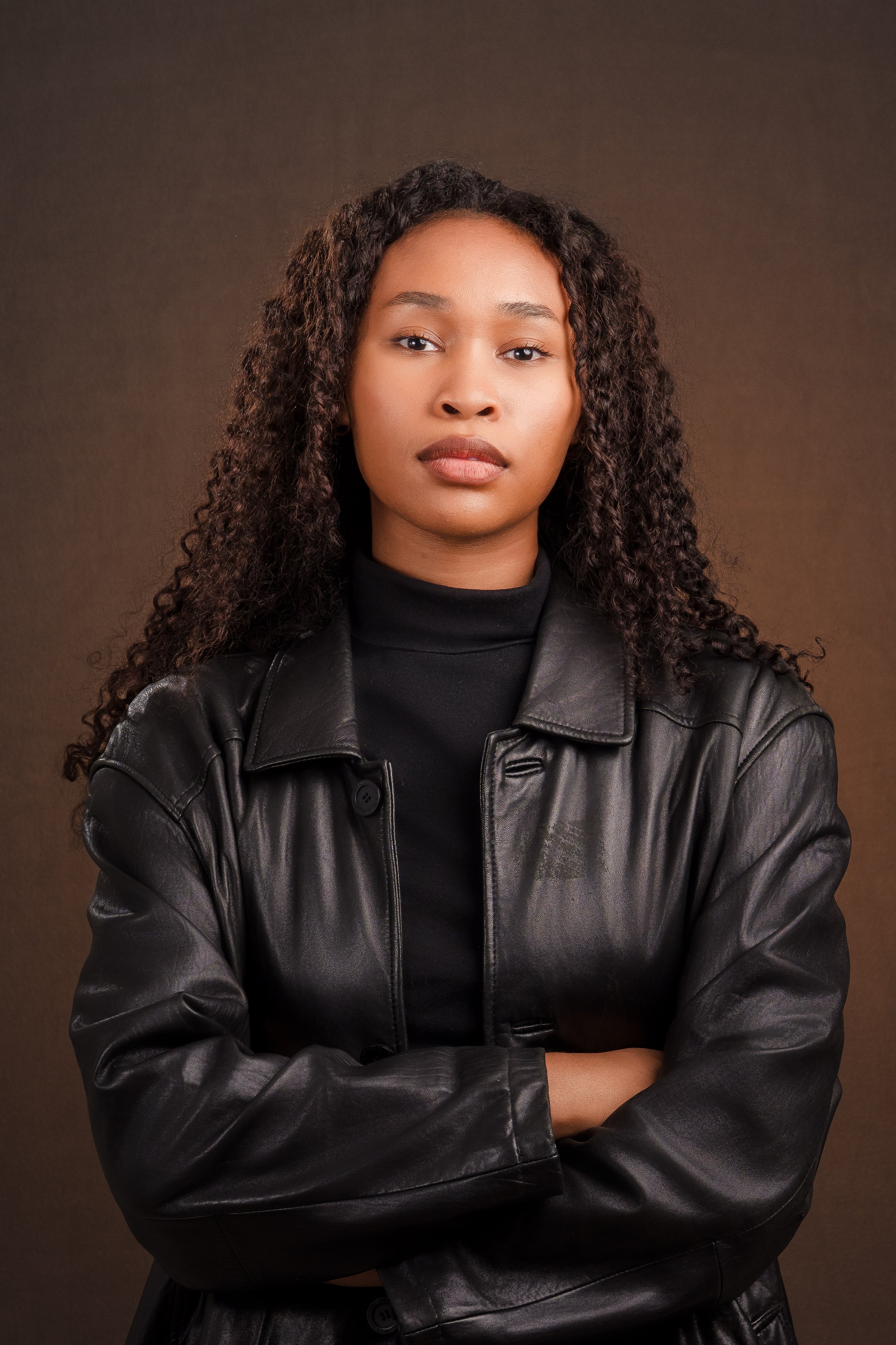 A woman with long curly hair, wearing a black turtleneck and a black leather jacket, standing with arms crossed against a plain brown background.