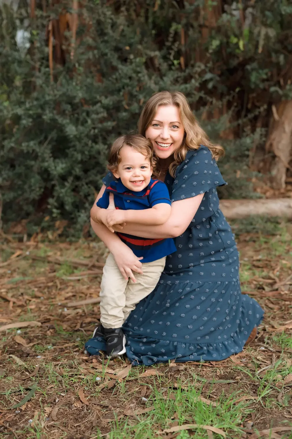 A mother and her young boy hugging and smiling outdoors in the wooded area of Gum Grove Seal Beach.