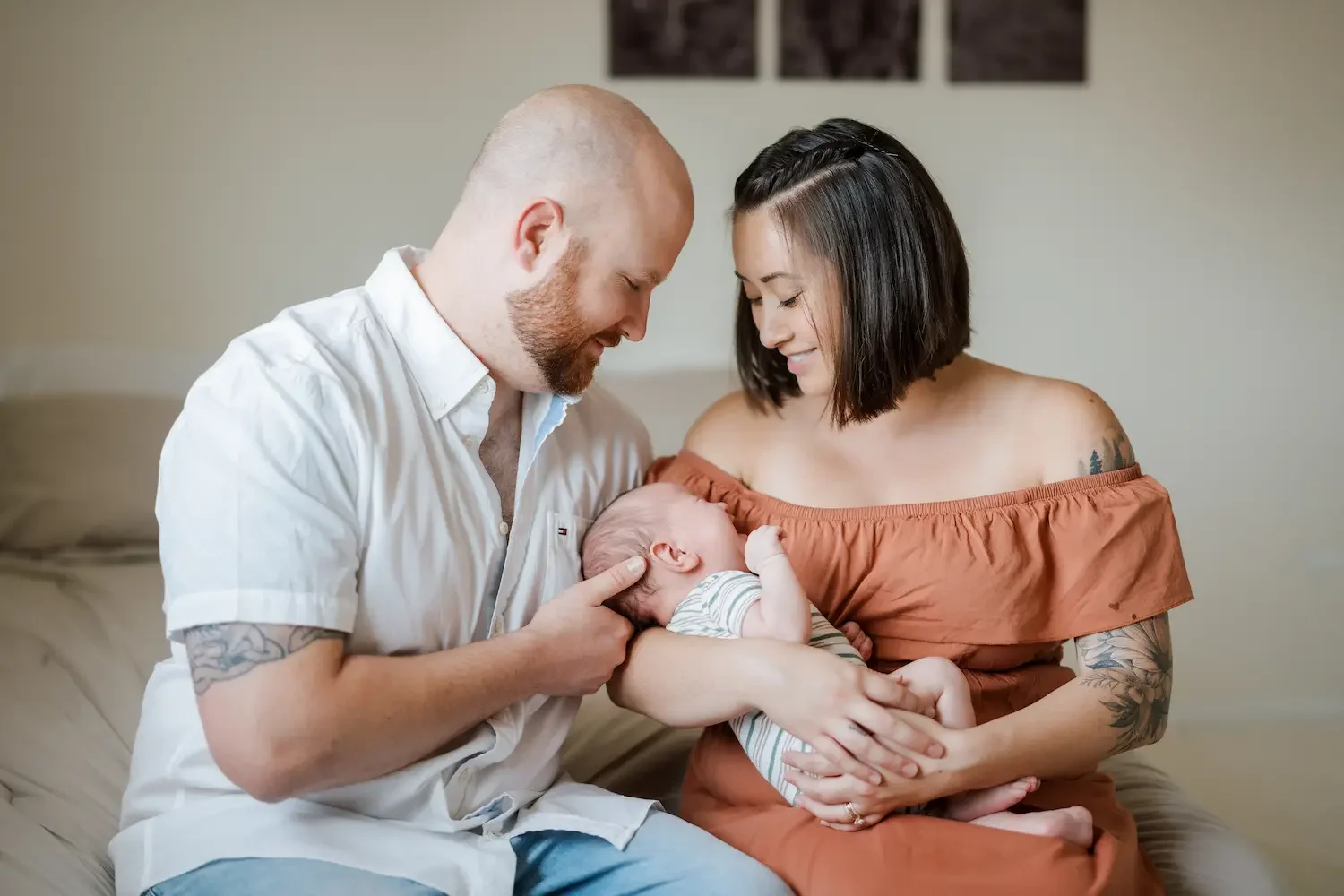 A couple holding a newborn baby, smiling and looking lovingly at the infant. The woman has tattoos and is wearing an off-shoulder dress, while the man wears a white shirt. They are sitting on a beige couch in a well-lit room.