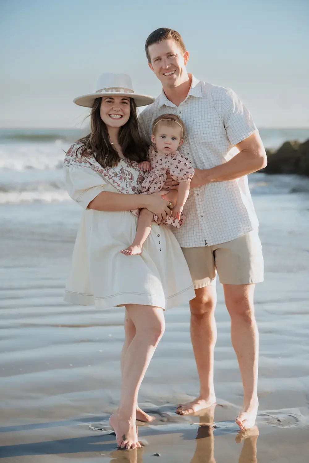 A family of three on the shoreline at Seal Beach, smiling and holding a baby girl. The woman wears a hat and a white dress, the man wears a white shirt and shorts, the baby wears a patterned dress. The ocean and sky are in the background.