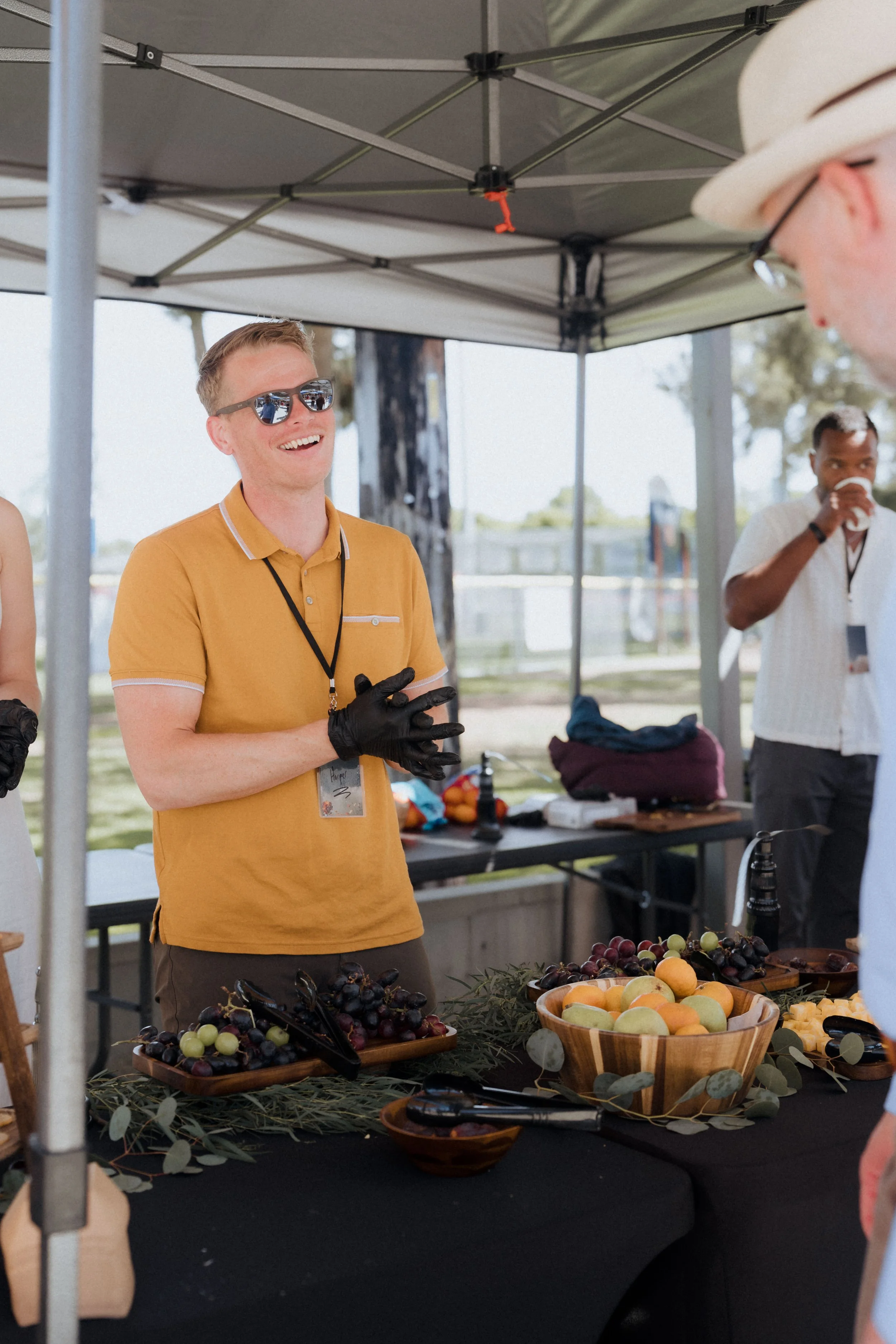 People at an outdoor fruit stand, with a young man in sunglasses and a yellow shirt smiling and wearing black gloves, surrounded by bowls of grapes and peaches.