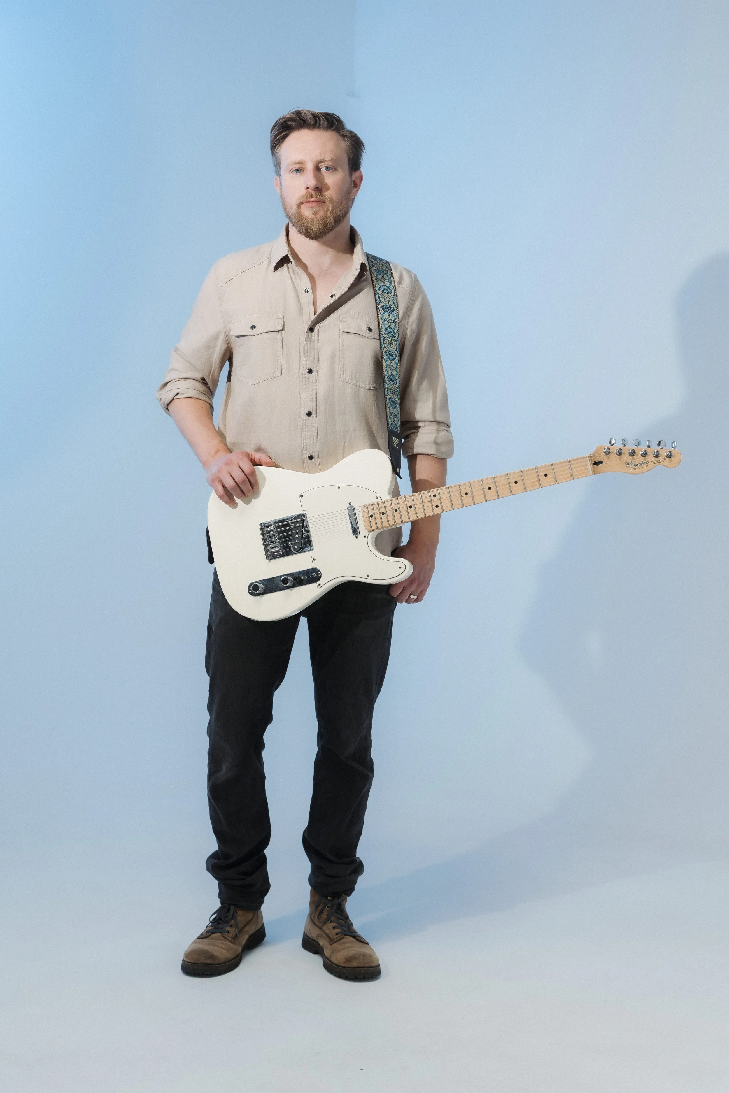 A man in beige shirt and black pants holding a white electric guitar against a plain blue background.