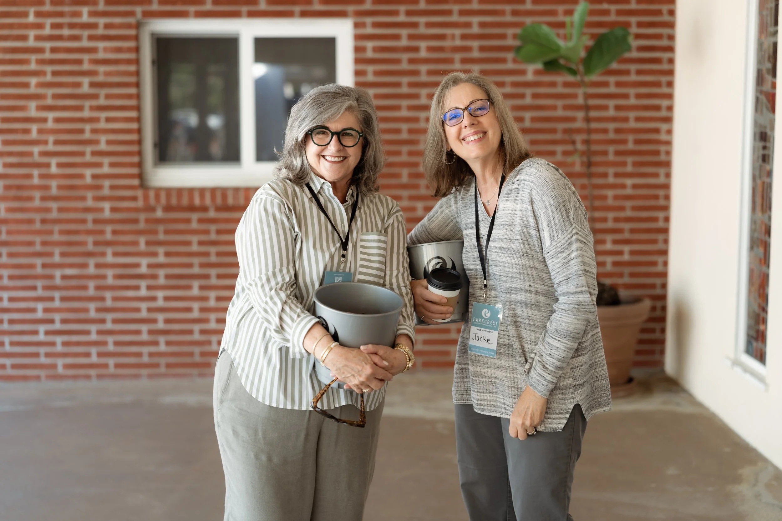 Two older women smiling and standing together outside in front of a brick wall, holding cups of coffee and plant pots, wearing conference badges.