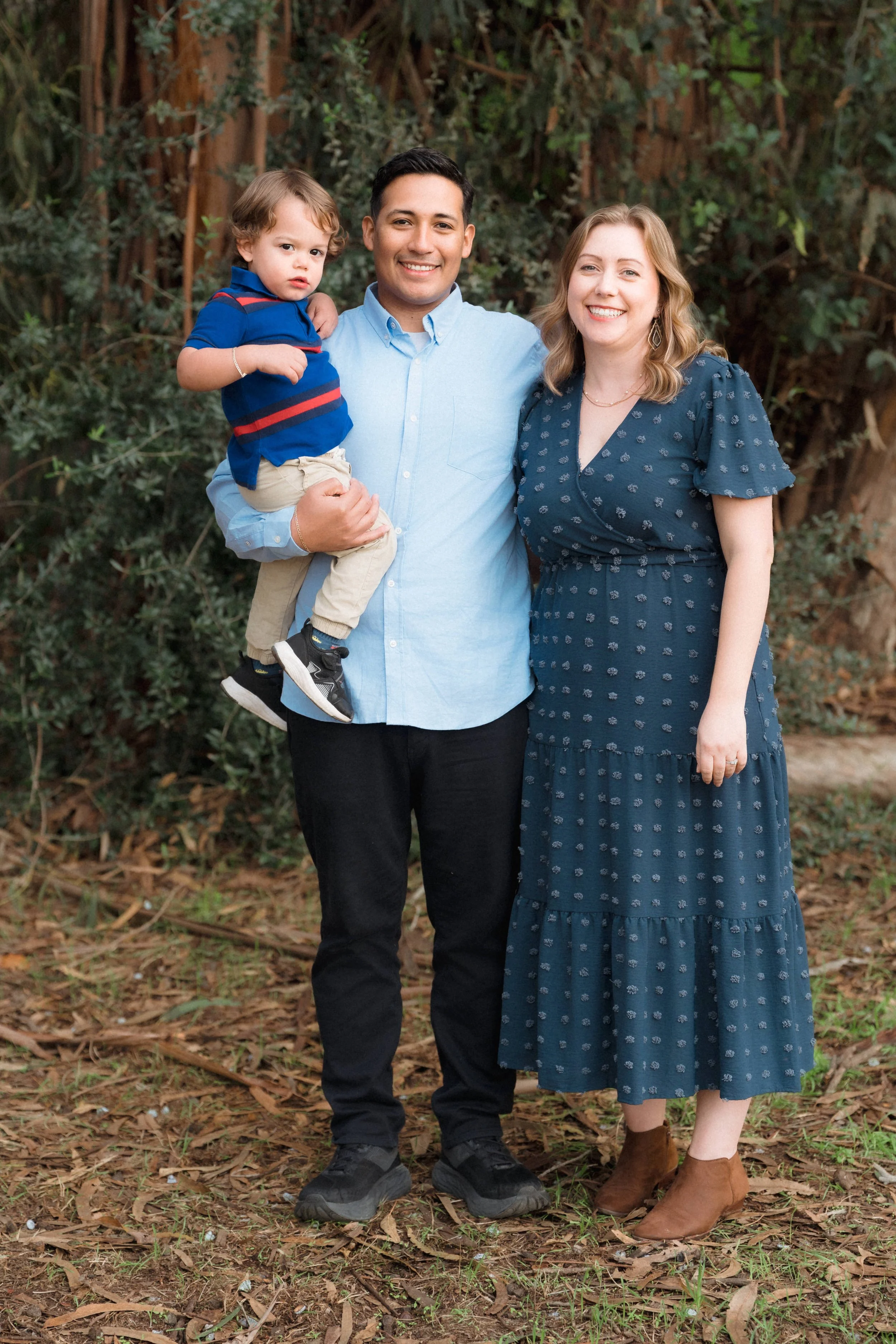 A smiling family of three standing outdoors with a wooded background. The man holds a young boy, and the woman wears a blue dress with brown ankle boots.