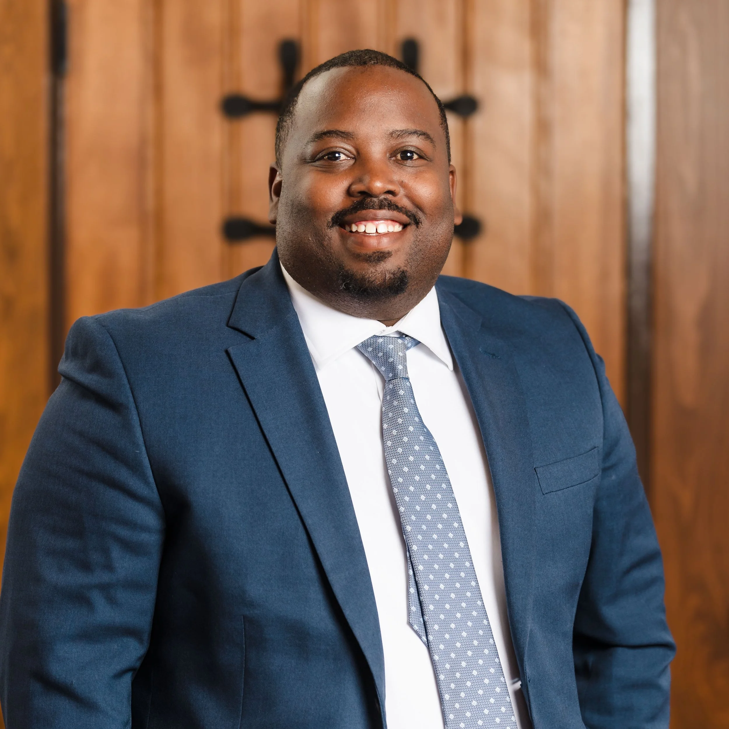 Close-up of a smiling African American man in a blue suit, white shirt, and patterned tie, with a wooden background.