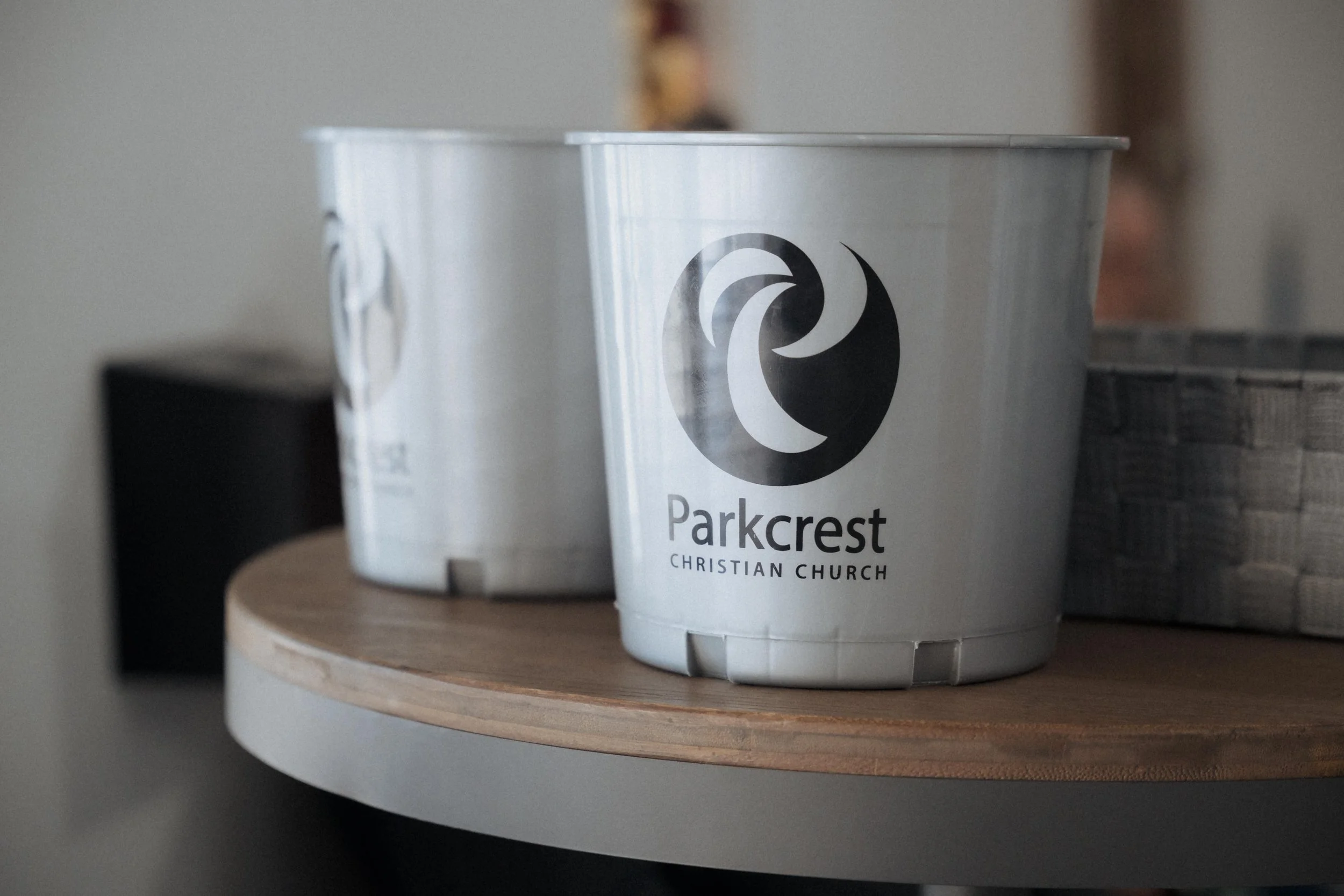 Two white plastic buckets with the logo of Parkcrest Christian Church, placed on a round wooden table.