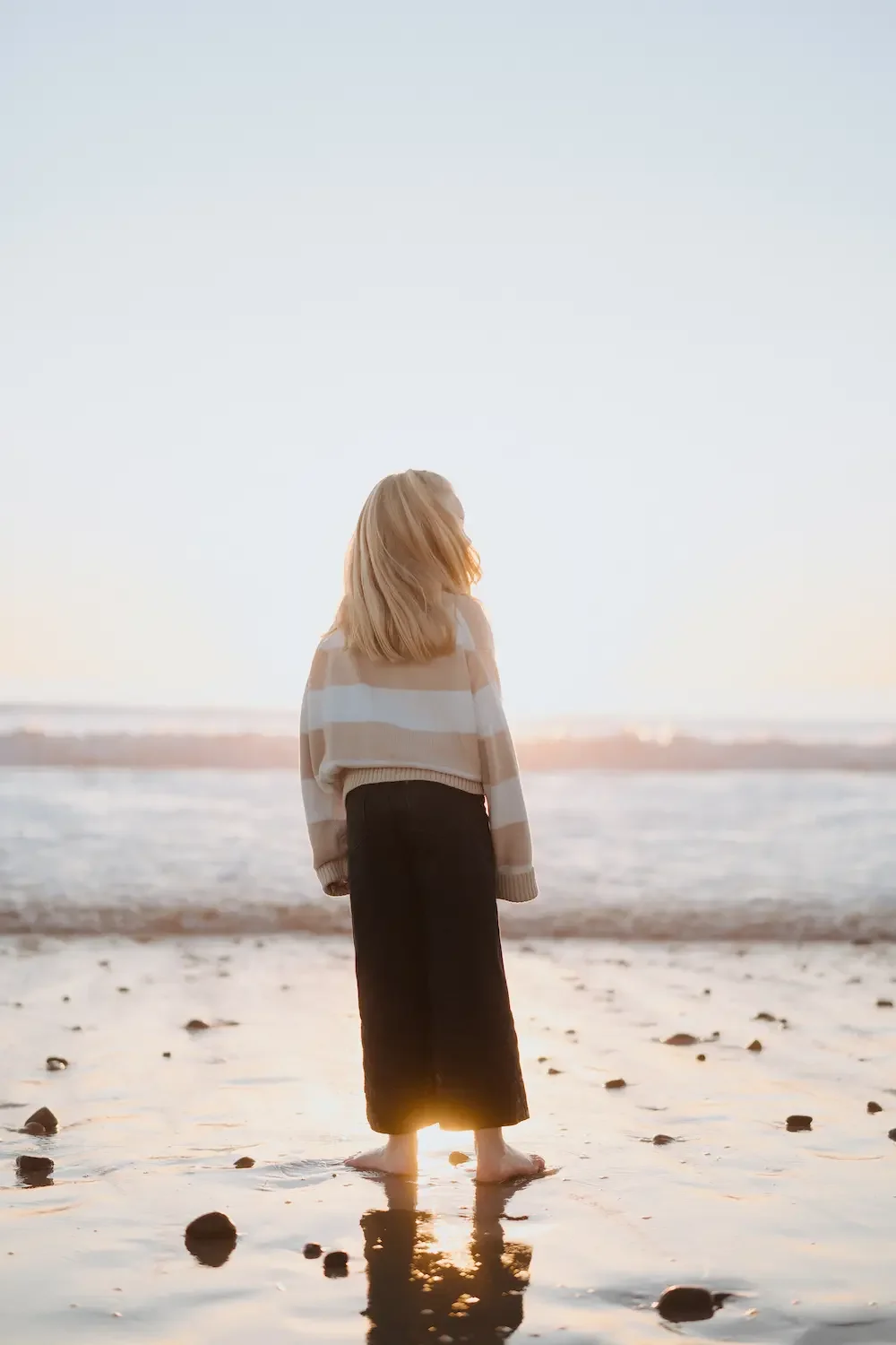 A young girl with blonde hair standing on a beach at sunset, facing the ocean and looking away from the camera.
