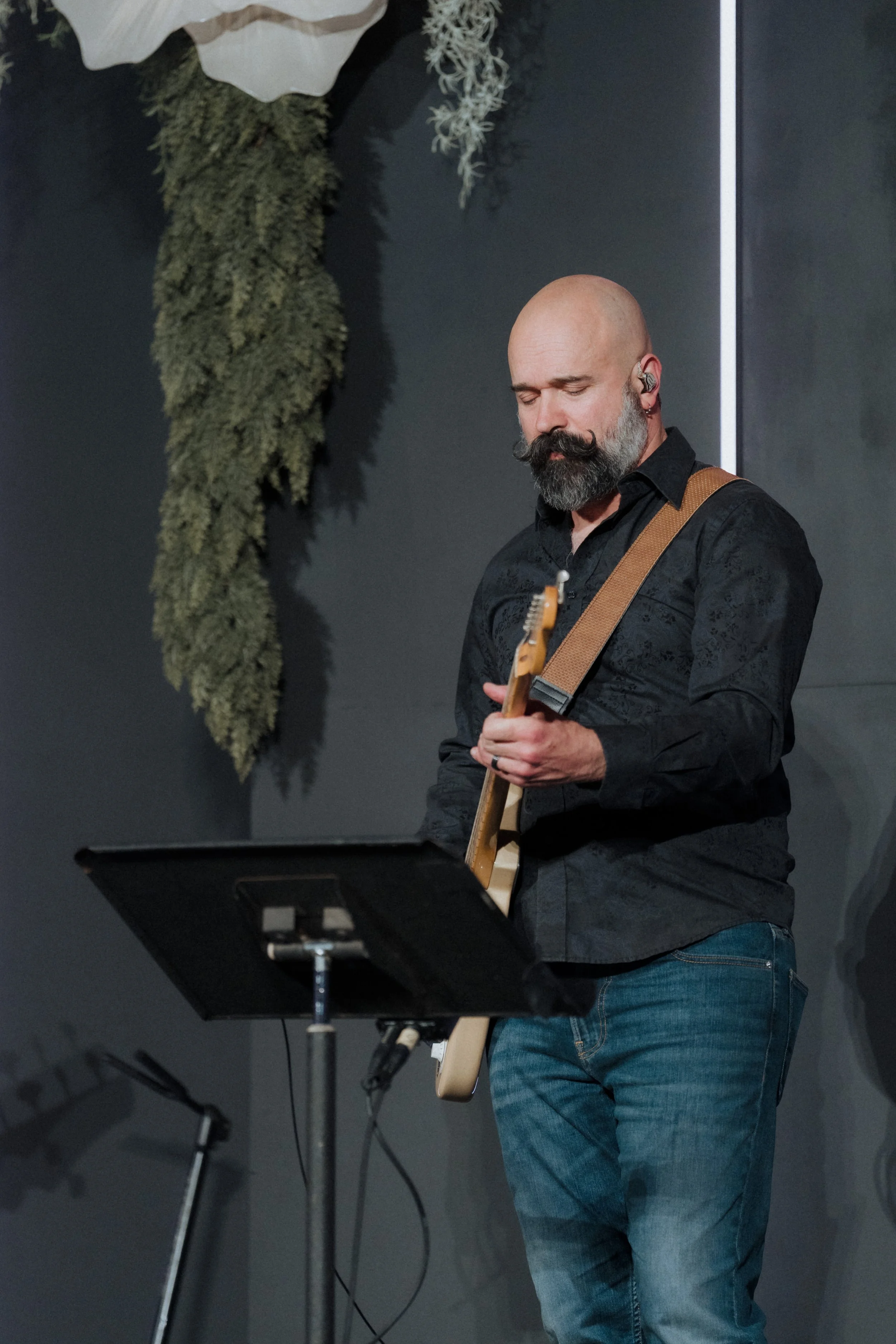 A man with a beard and mustache playing an electric guitar on stage, with a music stand in front of him, a black background, and some decorative greenery hanging on the wall.