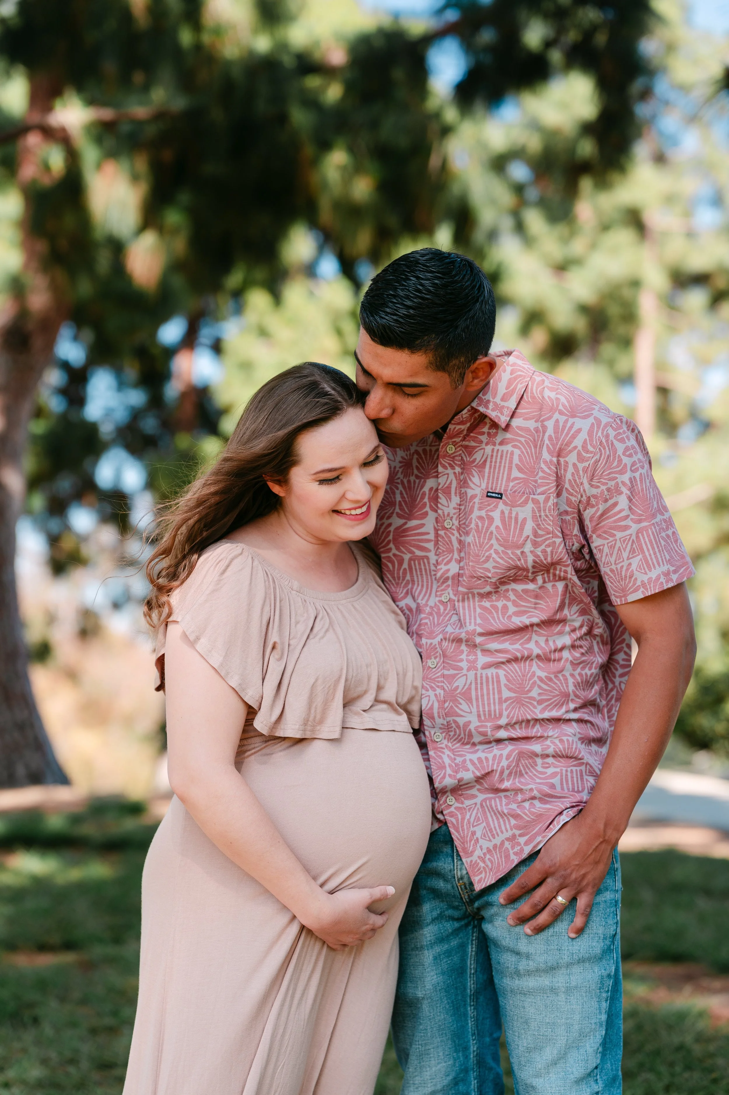 A pregnant woman in a beige dress smiling as a man kisses the top of her head in a park with trees in the background.