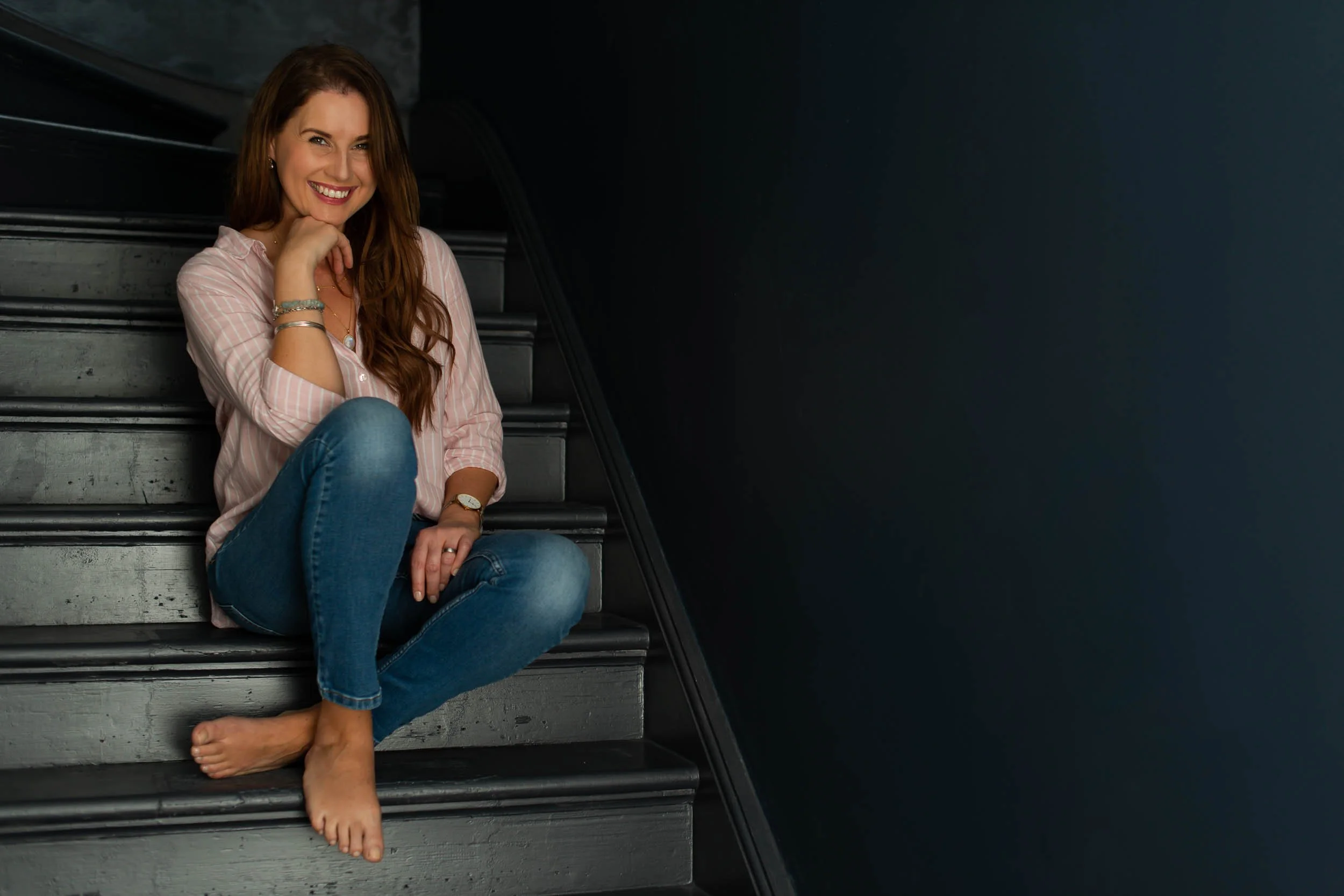 woman on stairs in jeans with a black backdrop smiling at the camera