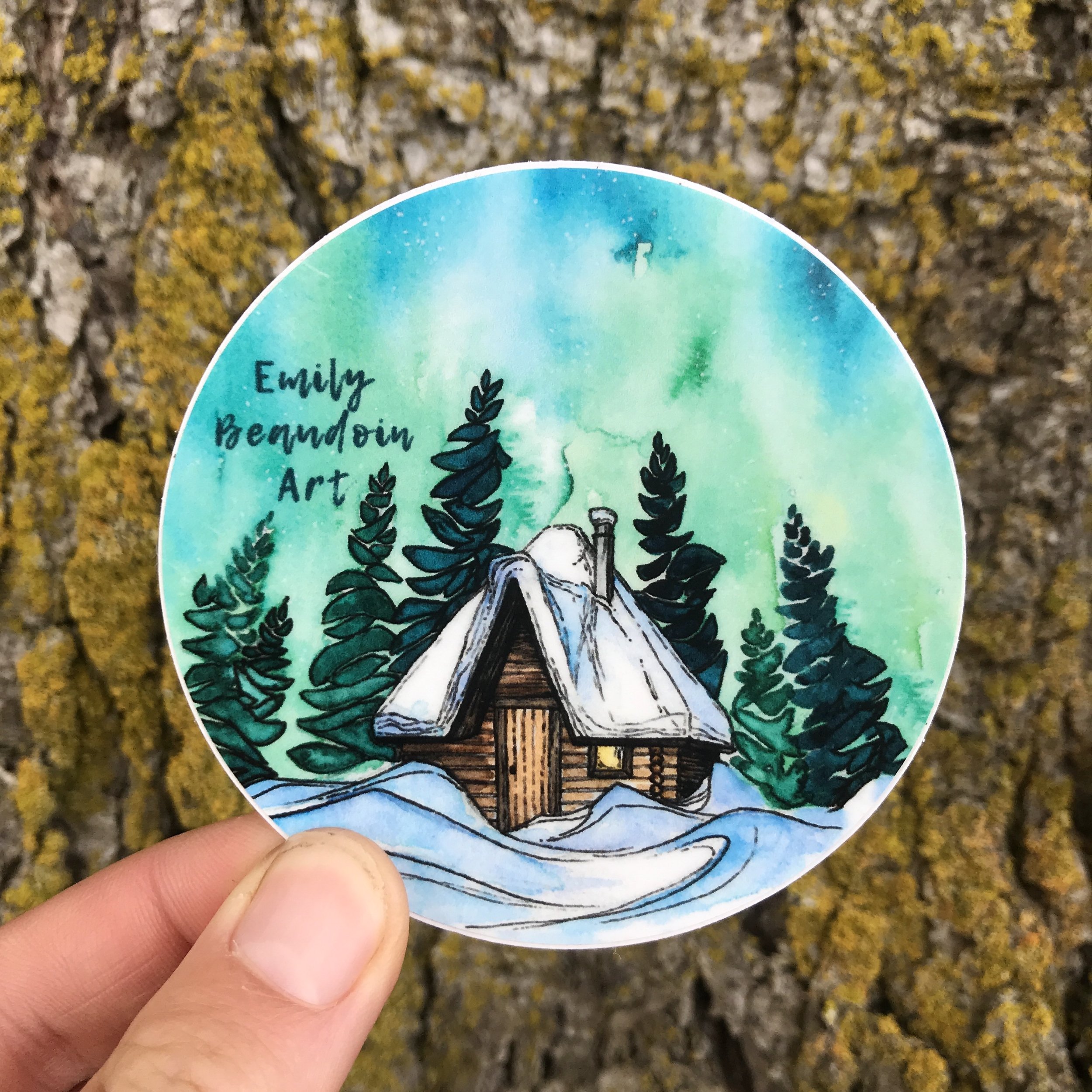 Artistic circular sticker featuring a snowy log cabin surrounded by pine trees and a colorful night sky with green and blue hues, with the text 'Emily Beaudoin Art' written on it.