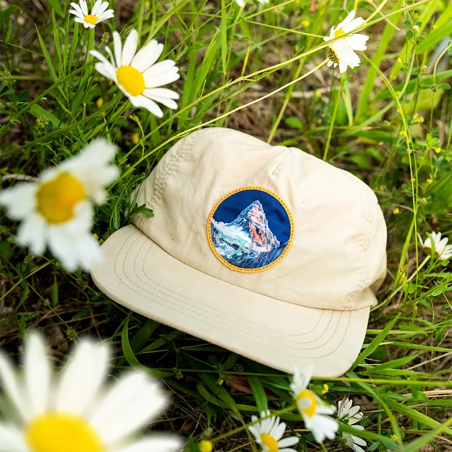 A beige baseball cap with a mountain patch lying on the ground surrounded by white daisies and green grass.