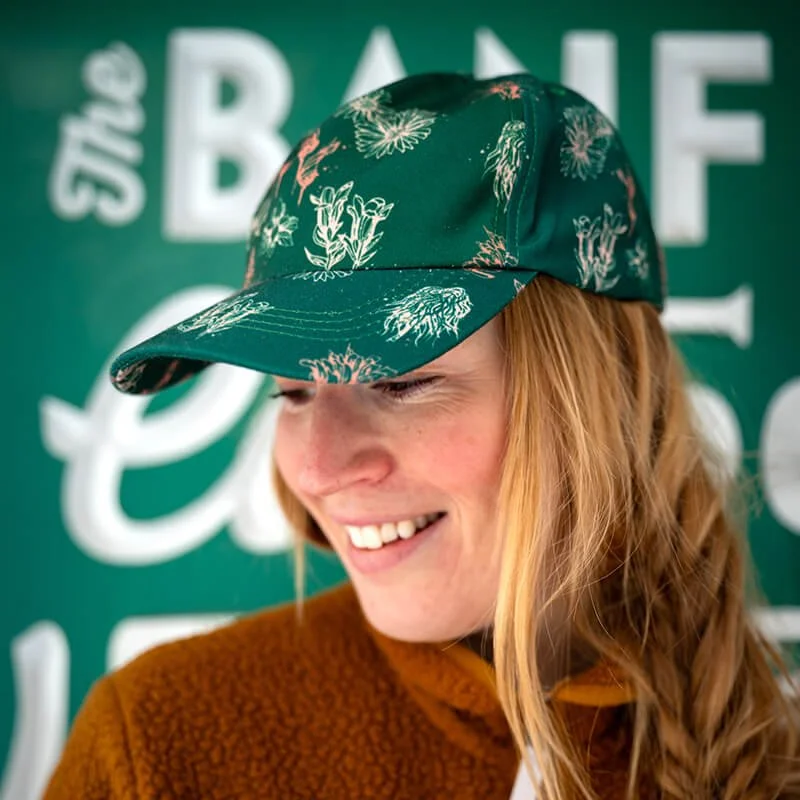 A smiling woman with long ginger hair wearing a green floral cap and a brown jacket, in front of a green background with white text.