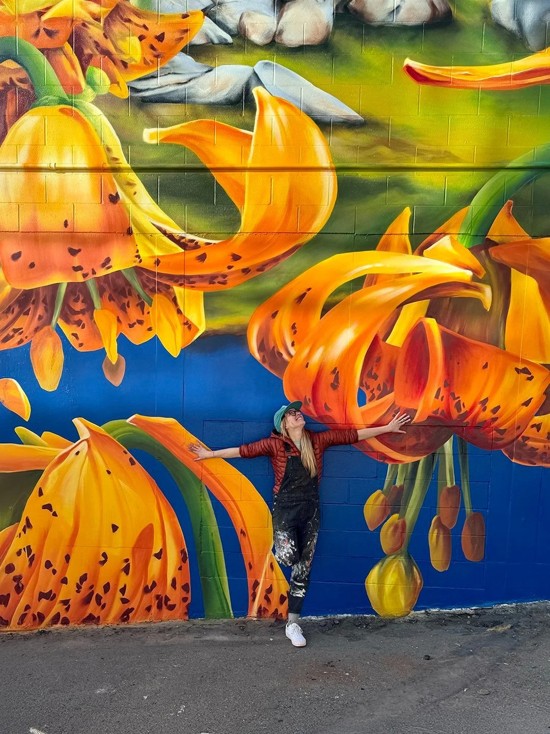A woman in overalls and a green hat smiling with arms outstretched, standing in front of a colorful mural of large orange and yellow lilies on a brick wall.