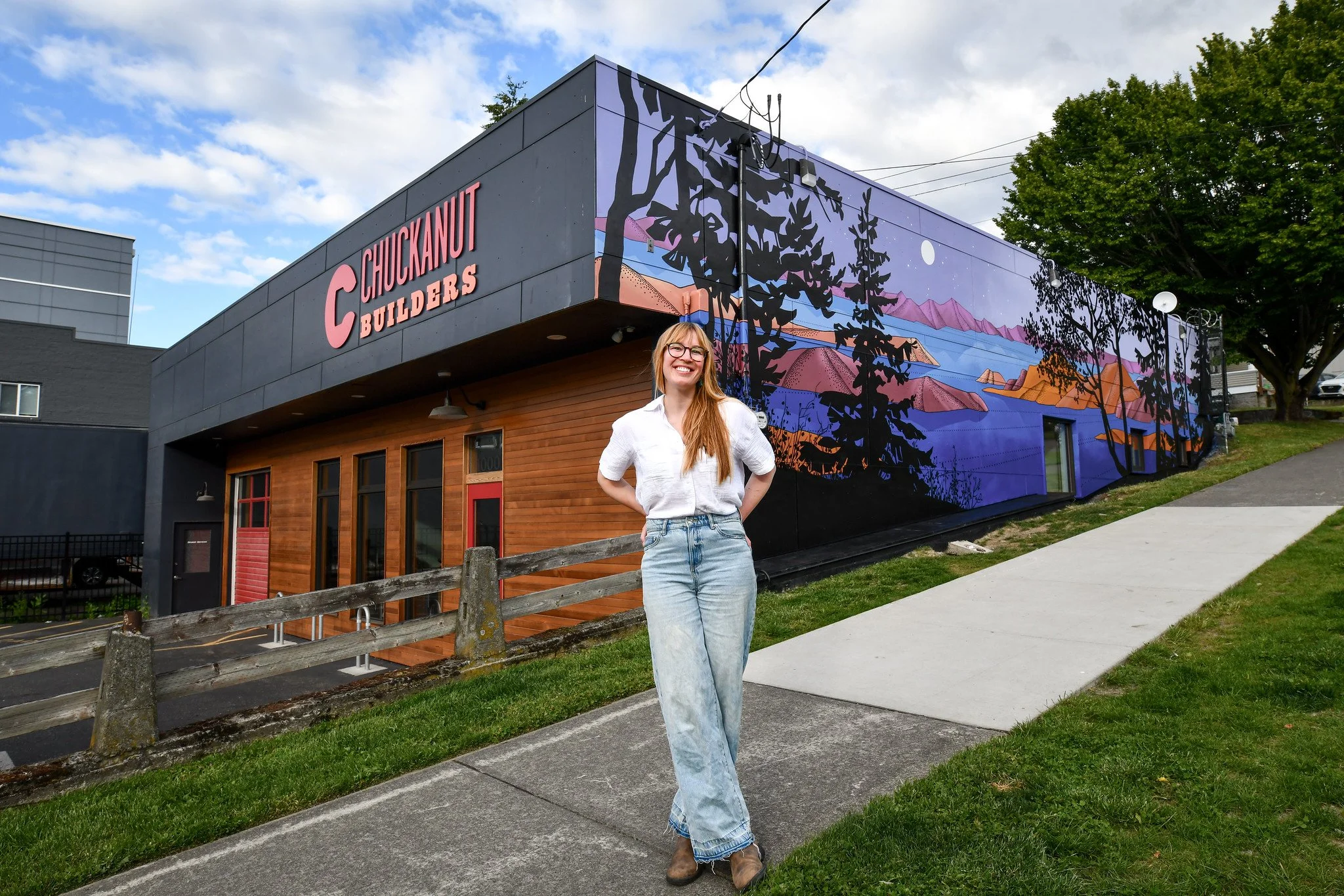 A woman with long red hair and glasses standing on a sidewalk in front of a building with a large mural. The building has a sign that reads 'Chuckanut Builders' and features a scenic landscape mural with trees, mountains, water, and a moon.