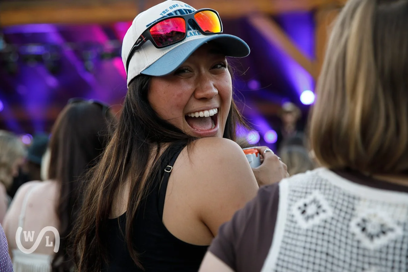 A woman with long brown hair, wearing a white and blue cap and sunglasses on her head, is smiling and looking at another person at a social event, with purple and blue lighting in the background.