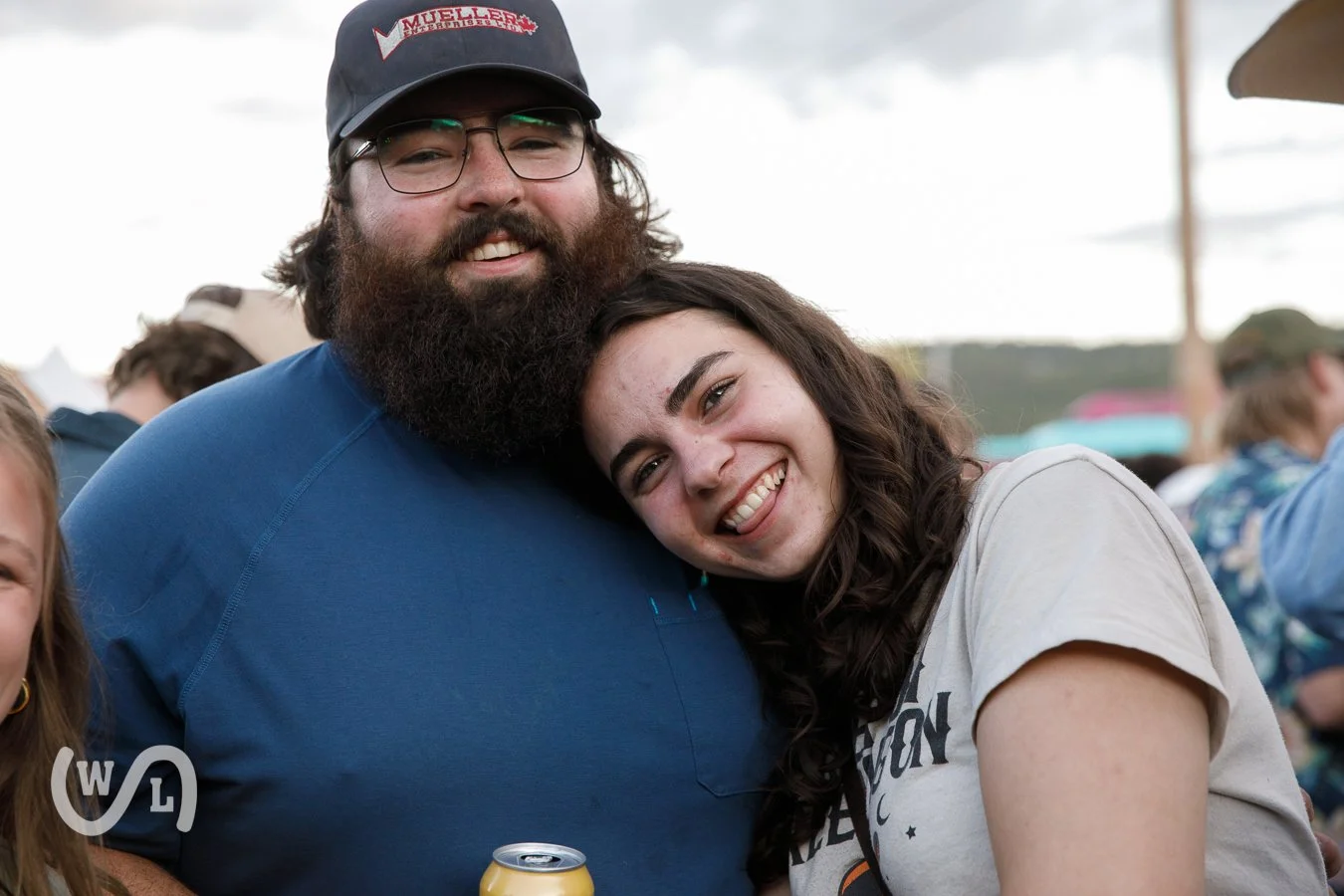 A man with glasses and a beard, wearing a baseball cap and blue shirt, hugging a young woman with long curly hair and a light-colored t-shirt, both smiling at an outdoor event.