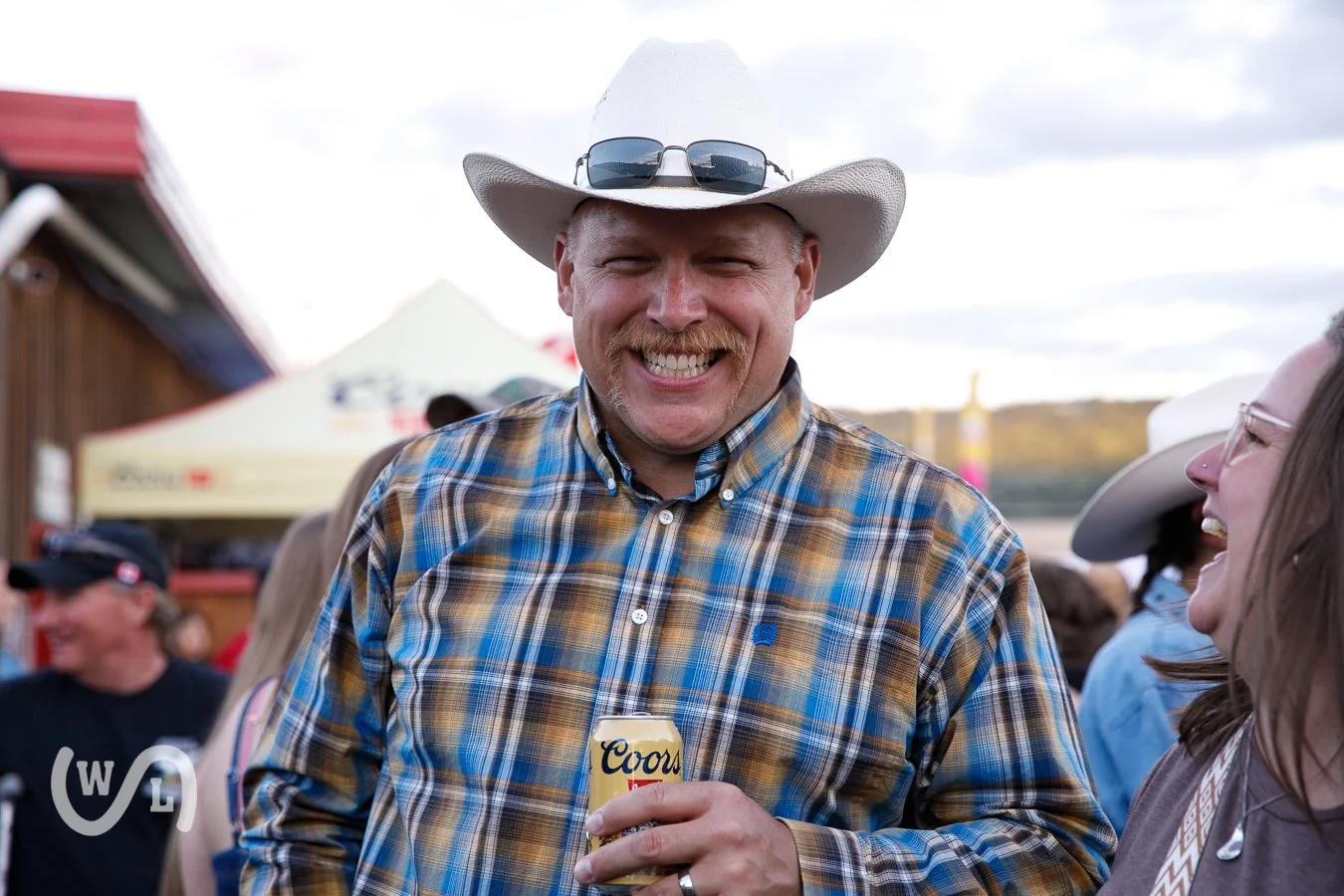 A man wearing a white cowboy hat, sunglasses on the brim, and a plaid shirt smiling and holding a Coors beer at an outdoor event, surrounded by other people laughing.