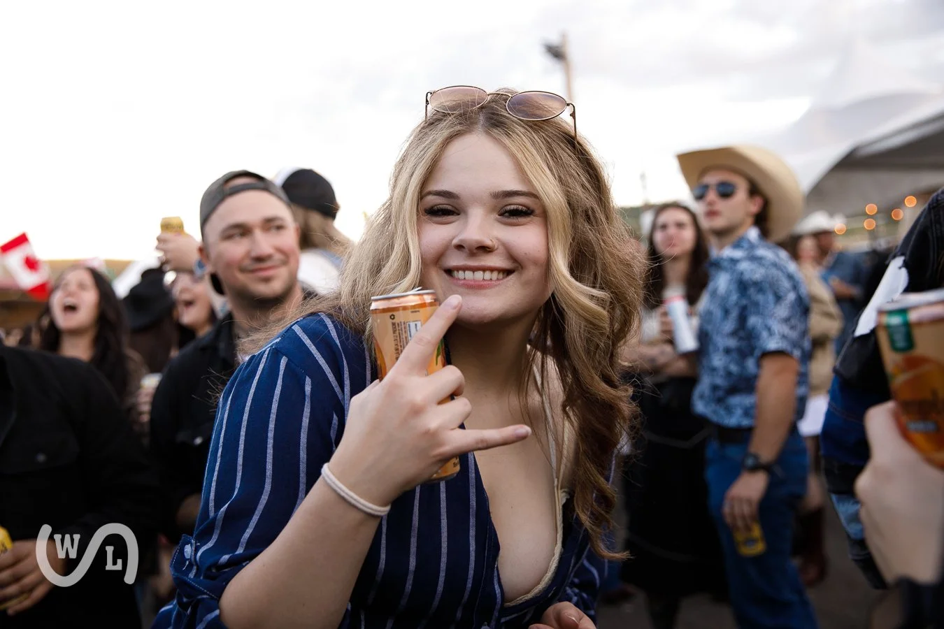 A young woman with wavy blonde hair smiling and holding a soda can at an outdoor event with a crowd in the background.