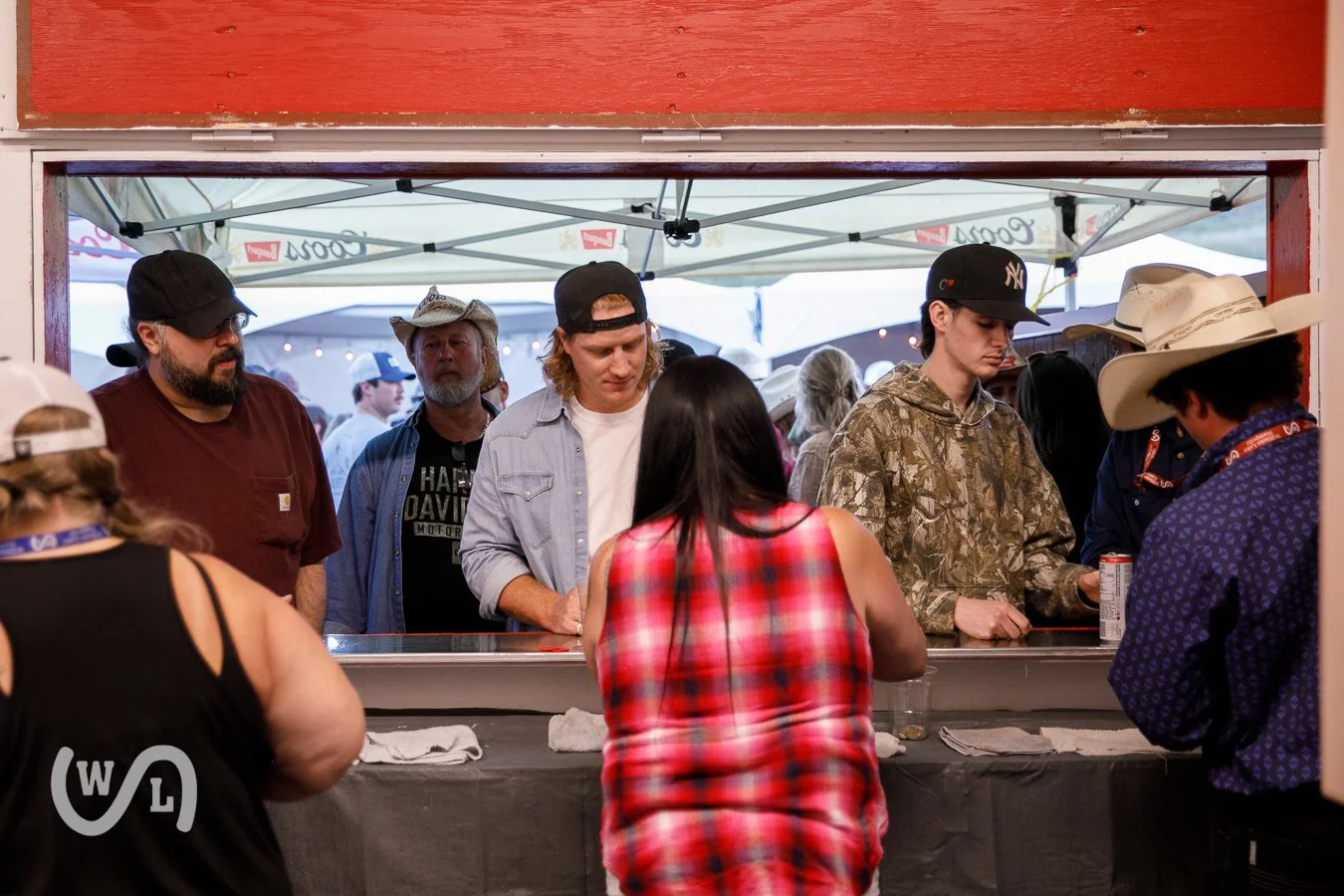 People at a food or drink stand in a festival setting, with a group of men and women waiting and serving behind the counter, some wearing hats and casual clothing.