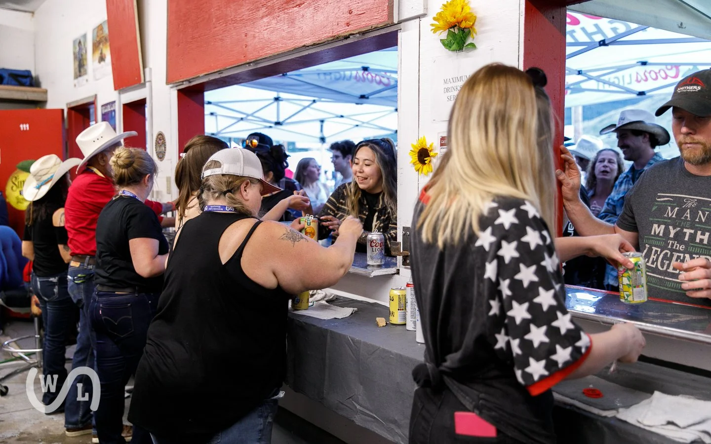 People in line at a crowded outdoor food stand, with some wearing cowboy hats, engaging in order transactions with the staff inside the stand, under blue and white tents.