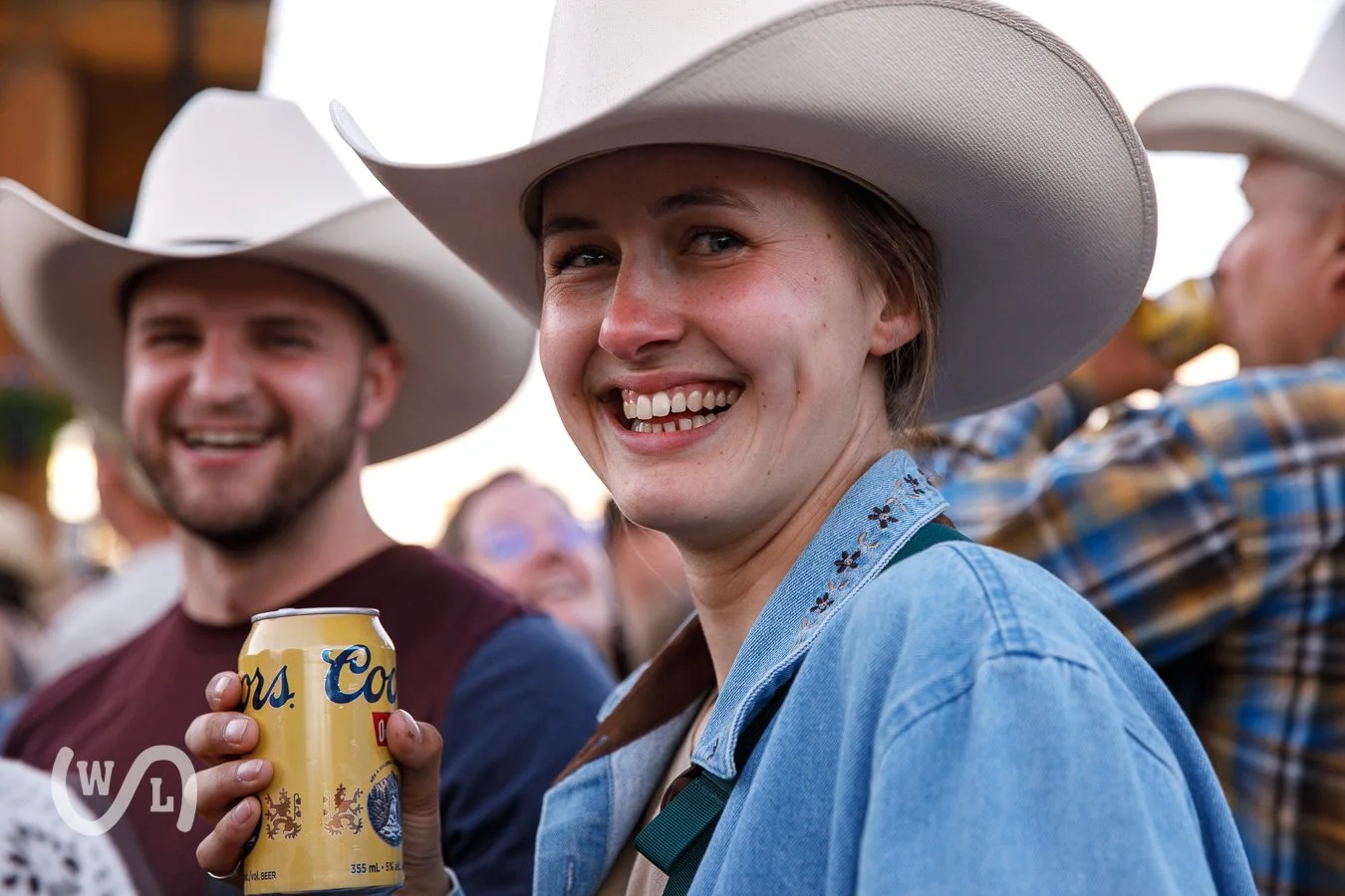 A woman smiling and holding a beer can at an outdoor event, wearing a cowboy hat and denim jacket, with other people in the background also wearing cowboy hats.