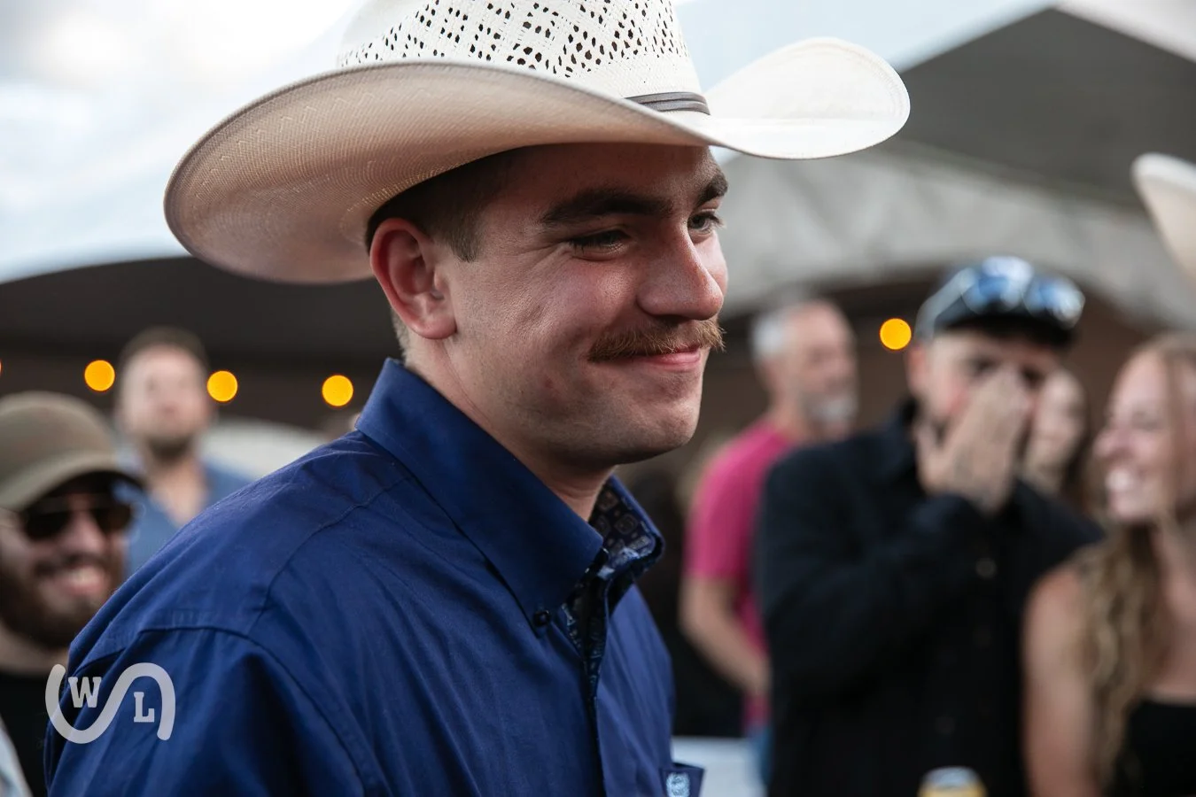 A man with a mustache wearing a cowboy hat and blue shirt, smiling at an outdoor gathering with blurred people in the background.