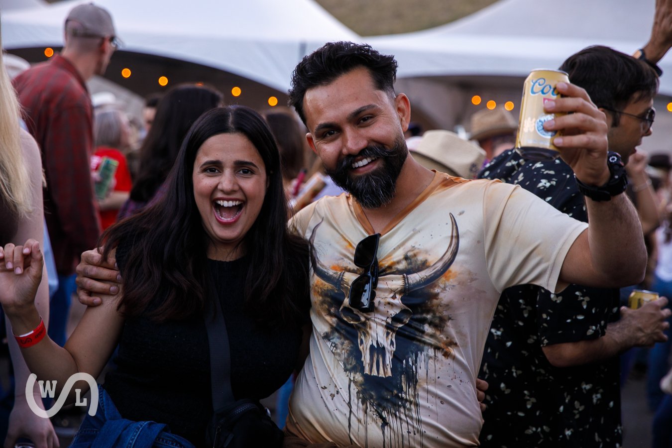 A smiling man and woman at a crowded outdoor event, with the man holding a can of Coors beer.