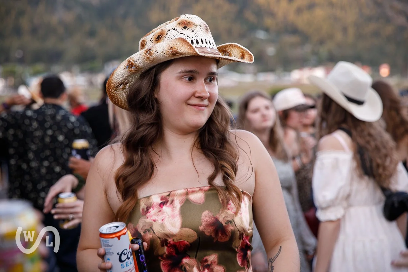 A young woman wearing a floral dress and a cowboy hat stands among a crowd at an outdoor event during the evening, holding a can of Coors soda.