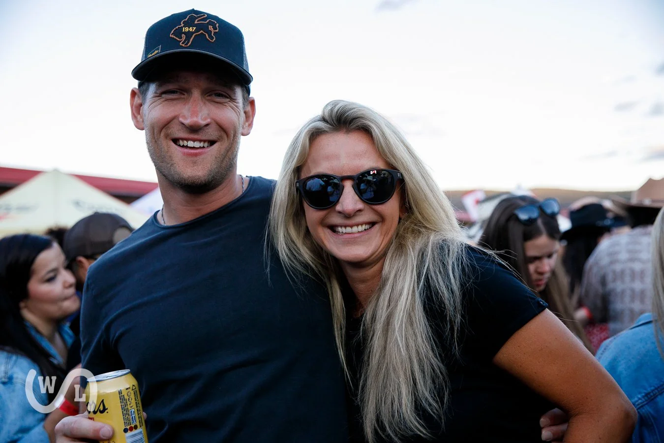 Smiling man and woman at an outdoor event, with tents in the background. The man is wearing a dark baseball cap and holding a can, the woman has long blonde hair and is wearing sunglasses.