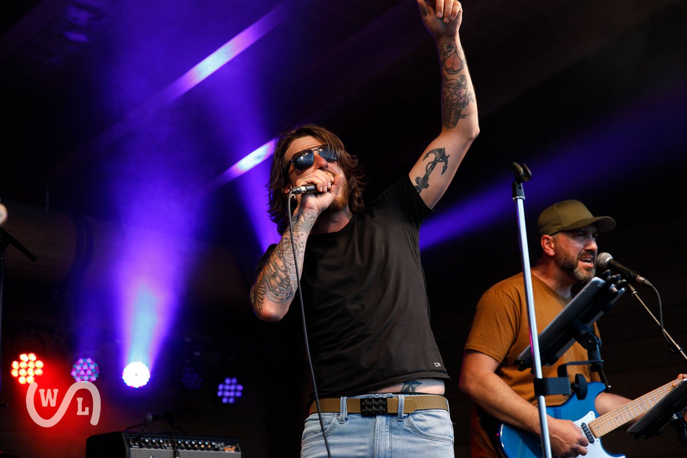 Musician with tattooed arms, wearing sunglasses, black t-shirt, and jeans, singing into microphone with one arm raised on stage, another musician playing guitar nearby, colorful stage lights in background.