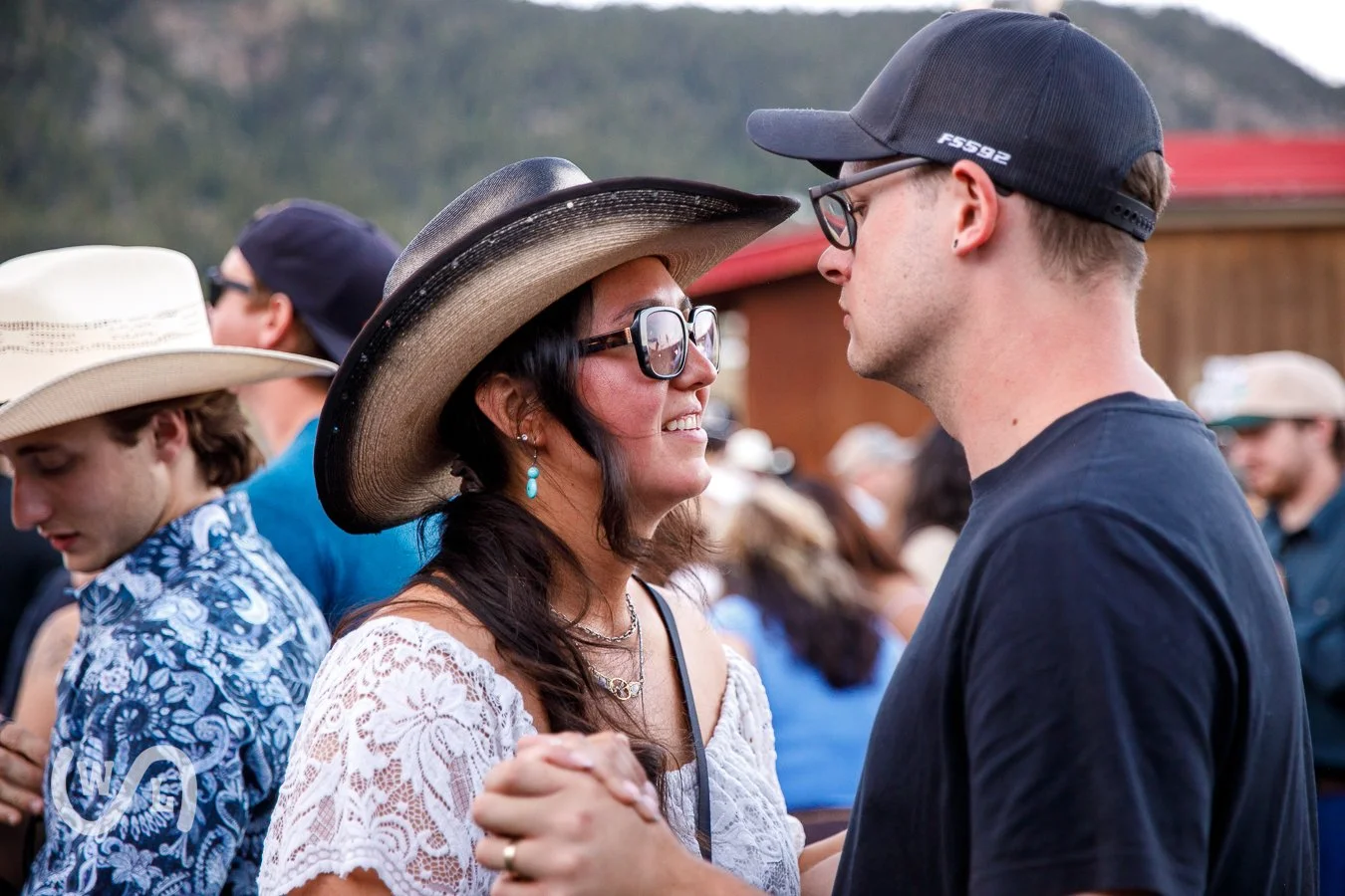 A woman and a young man in a black t-shirt and cap face each other closely, holding hands and smiling at each other, at an outdoor event with other people in the background.