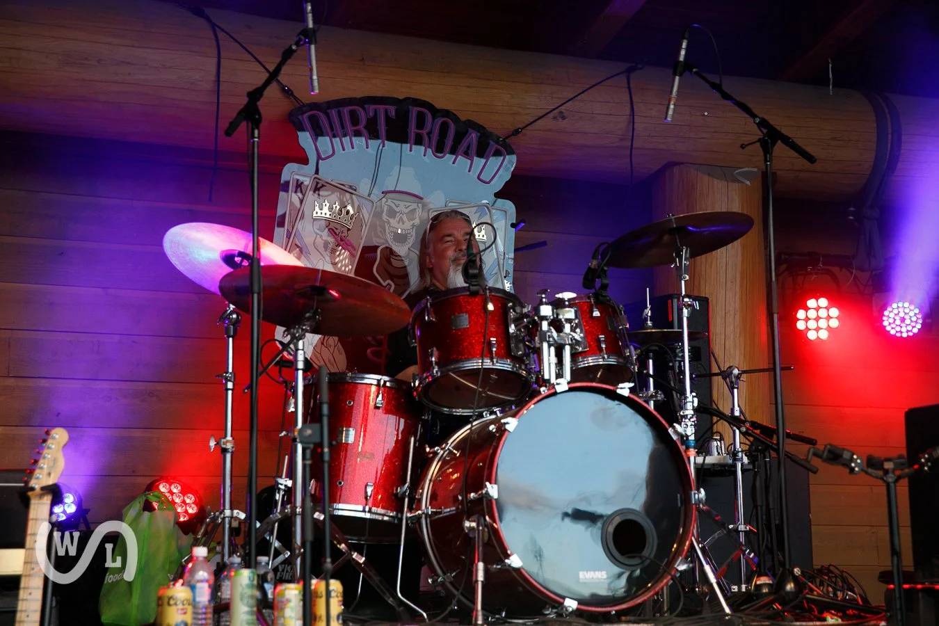 A man playing a red drum set on stage with a 'Dirt Road' band banner behind him and various stage lights around.