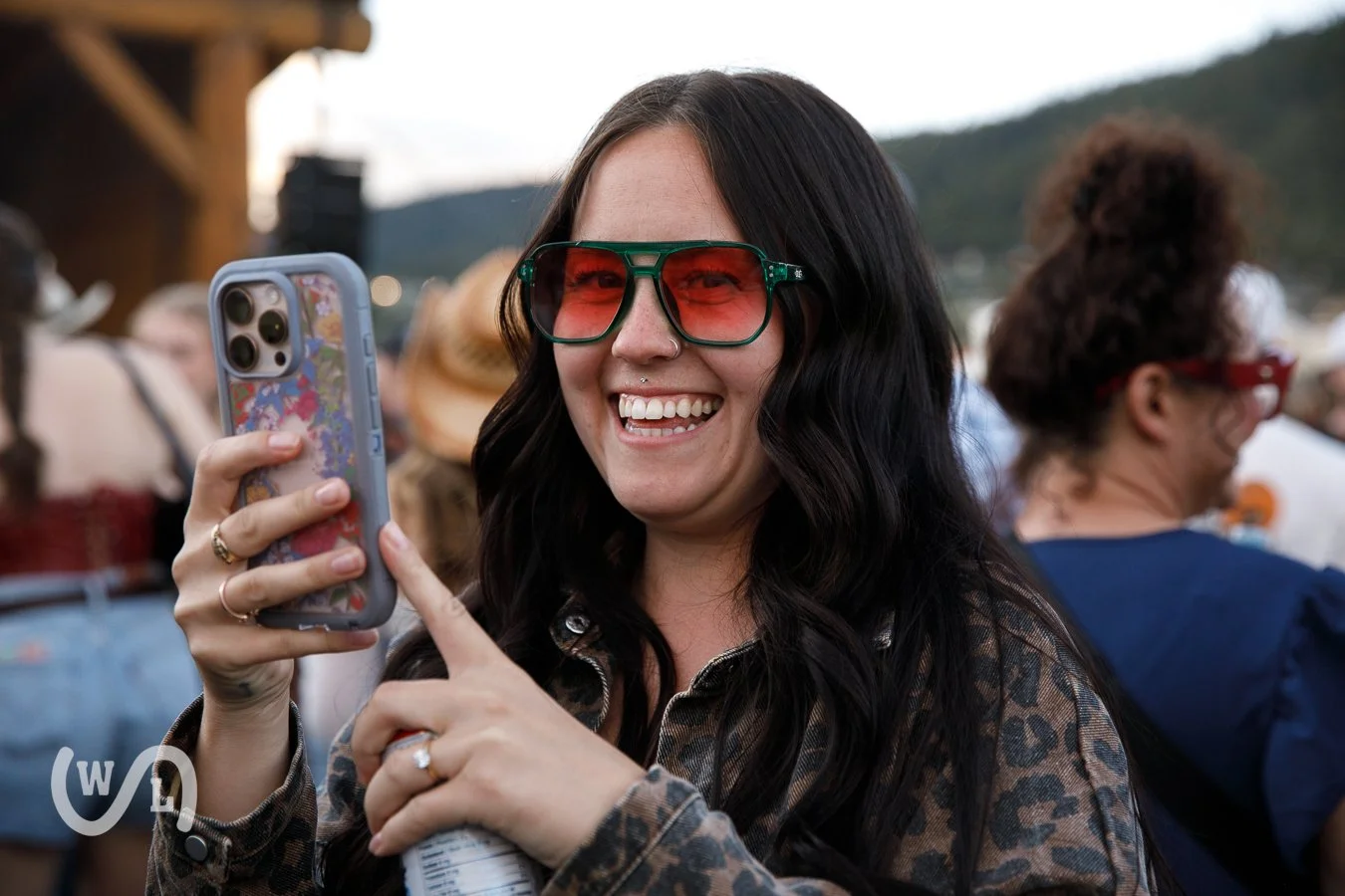 A woman with long black hair wearing red-tinted sunglasses smiling and taking a photo with her phone at an outdoor event.