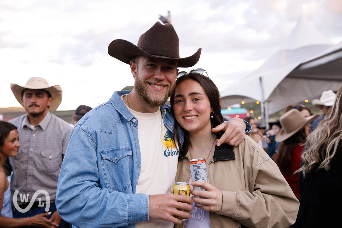 Two smiling young adults, a man and a woman, holding drinks at an outdoor event with people and tents in the background, both wearing cowboy hats.