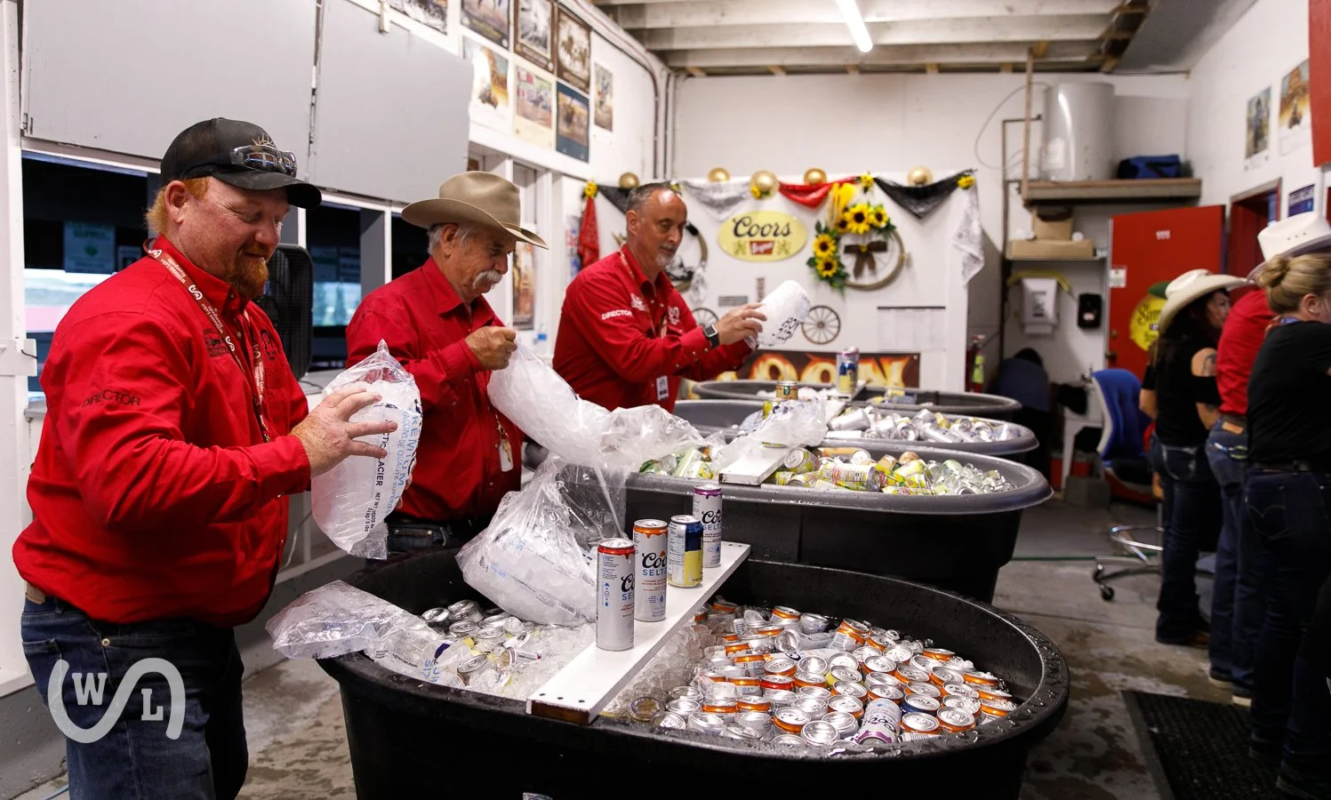 Four men in red shirts working at a beer bottle recycling station, sorting and crushing empty cans in large black bins inside a room decorated with posters, sunflower decorations, and a banner that says "Coors".