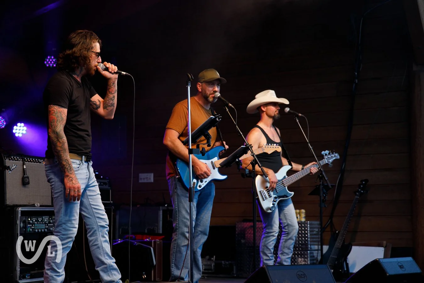 Three musicians performing on stage with electric guitars, one wearing a cowboy hat, another in a cap, and the third with tattoos, in a dimly lit setting with purple stage lighting.