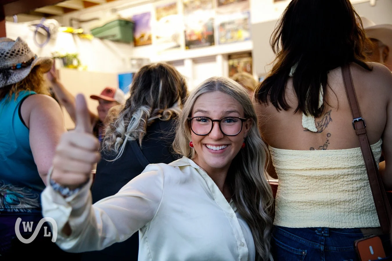A smiling woman with glasses and long wavy hair in a white shirt gives a thumbs-up at a crowded indoor gathering.