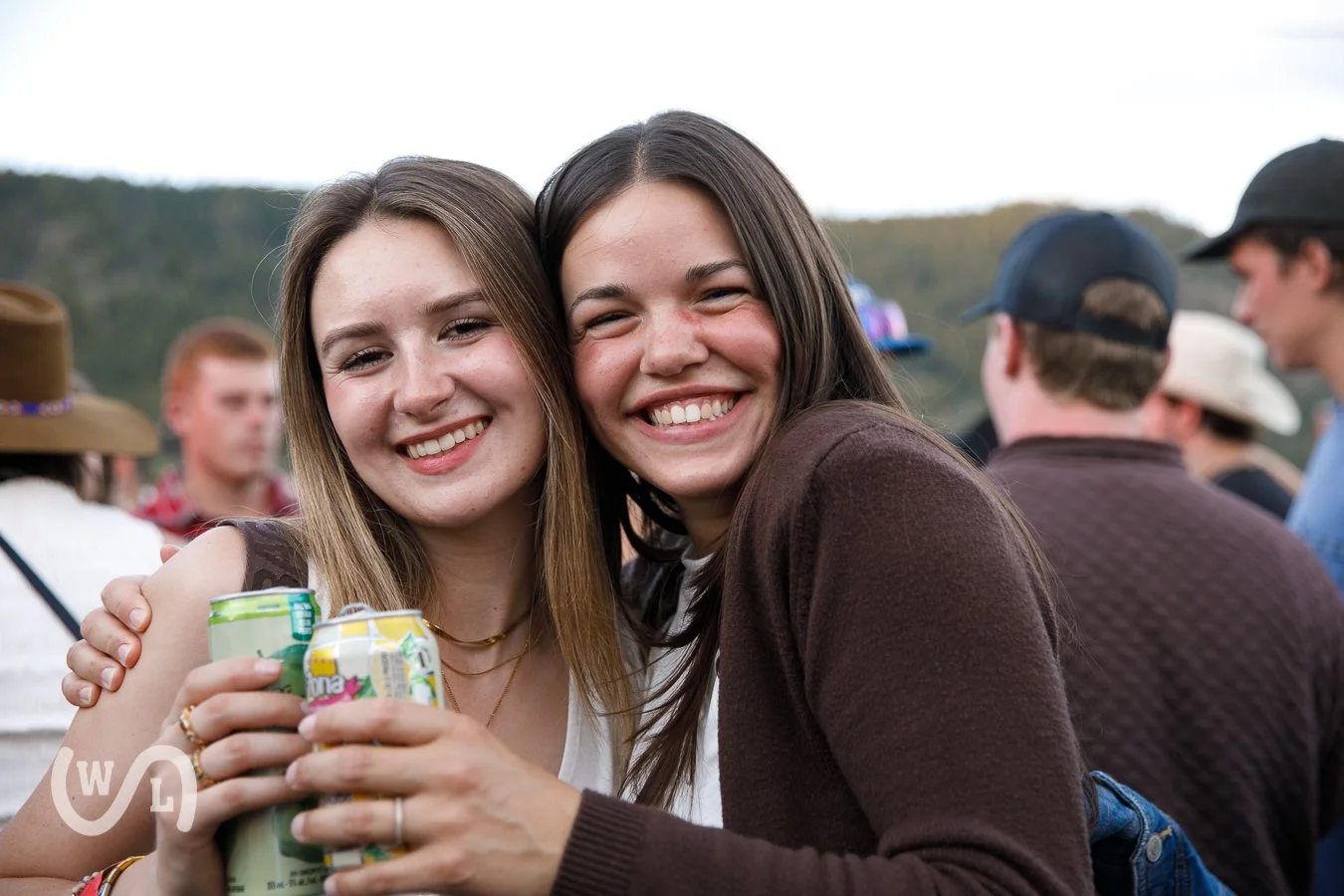 Two young women smiling and embracing at an outdoor gathering, holding canned drinks, with other people and a natural landscape in the background.