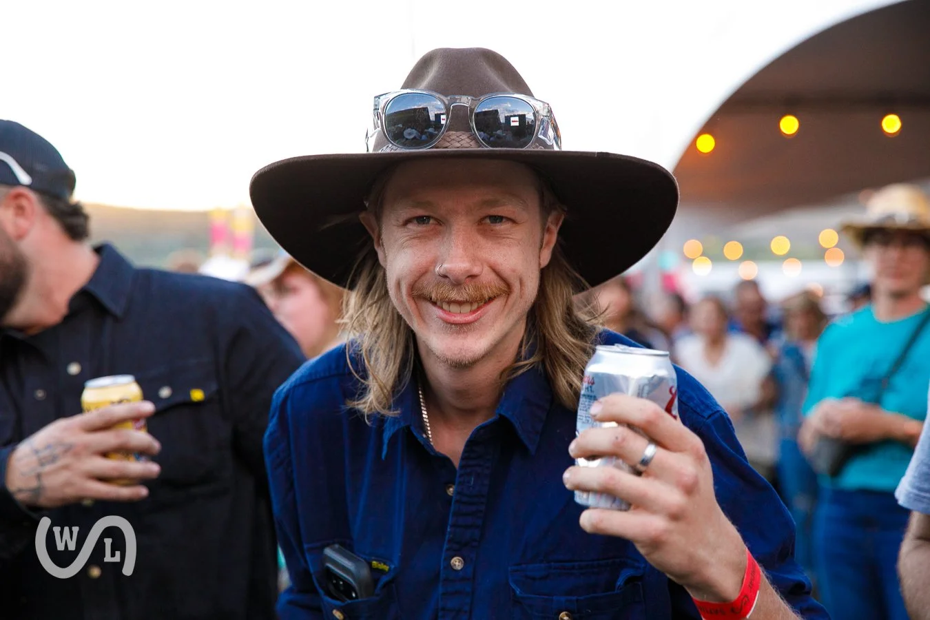 A smiling man wearing a large hat, sunglasses on the hat, and a blue shirt, holding a can and surrounded by people at an outdoor event during evening.