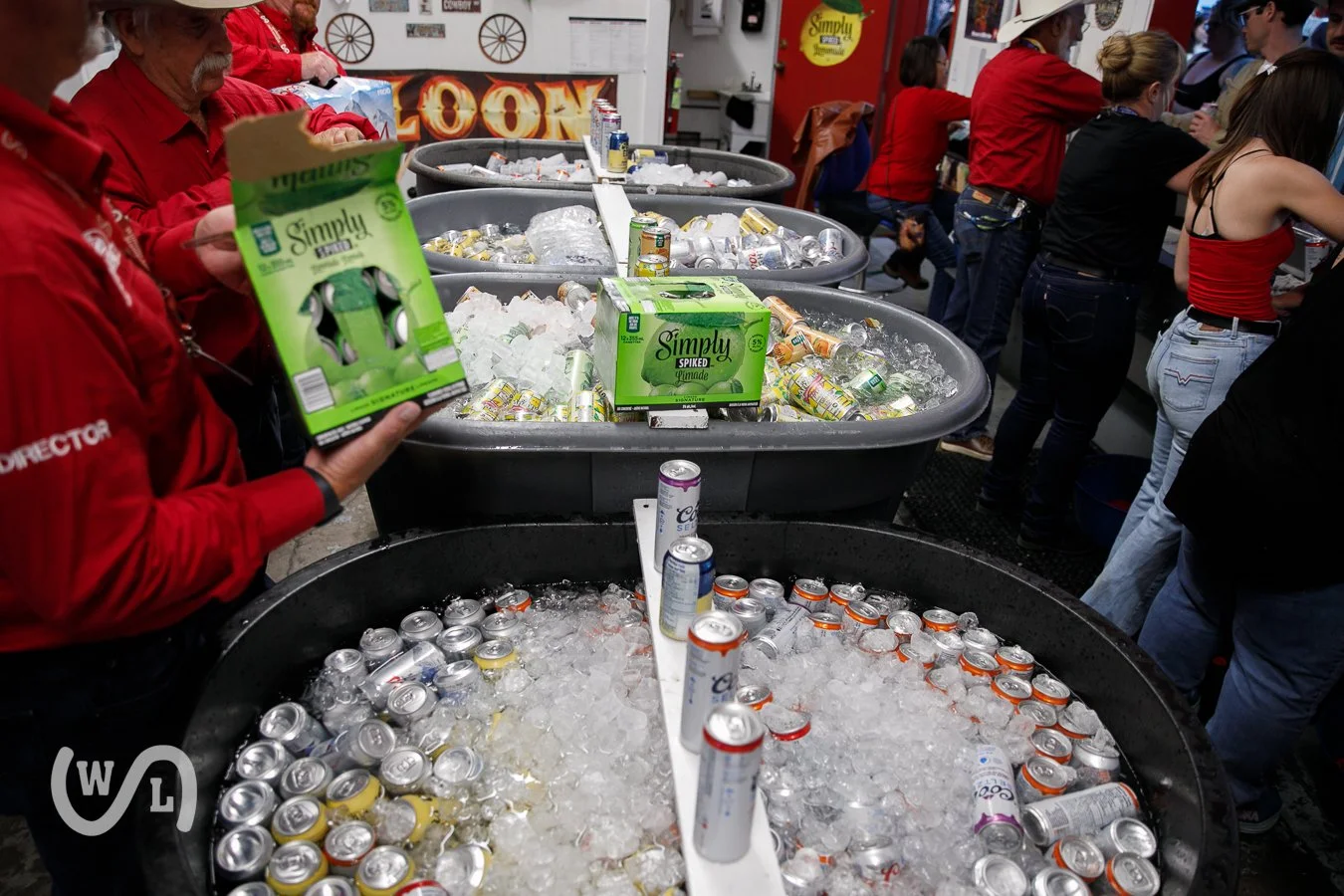 People at a bar or club with large containers of ice filled with cans of beer and hard seltzers. Some individuals are serving drinks while others wait in line. The setting appears lively and festive, with a red, white, and black color scheme and vari