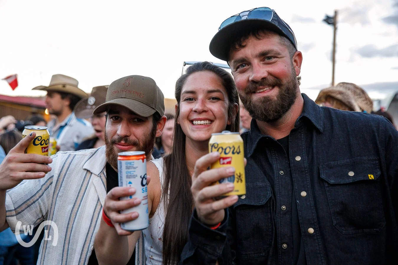 Group of three friends smiling and holding cans of Coors beer at outdoor event.