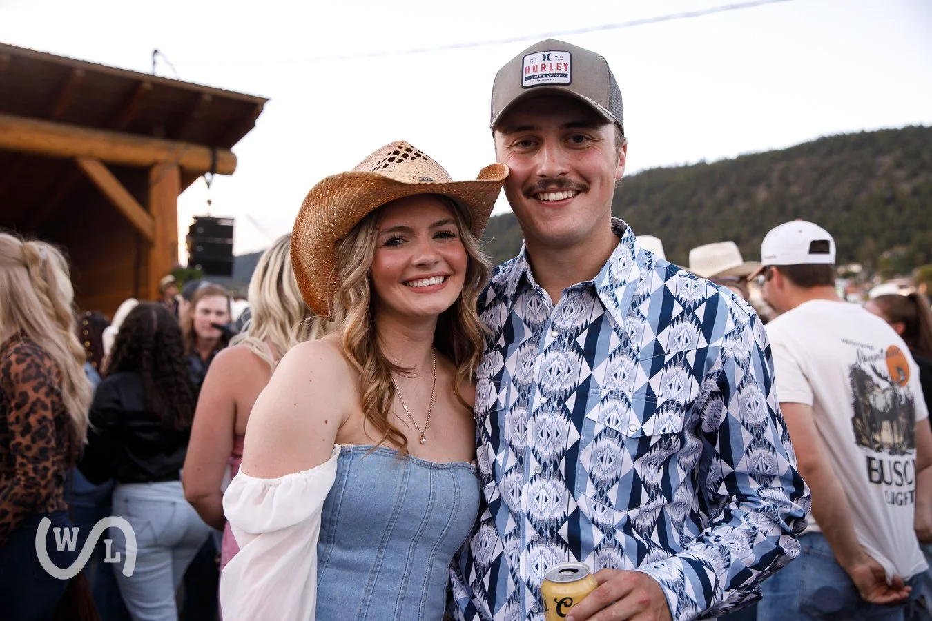 A smiling young woman in a cowboy hat and off-shoulder top standing next to a smiling man in a patterned shirt and baseball cap, holding a canned drink, at an outdoor event with a crowd and mountains in the background.