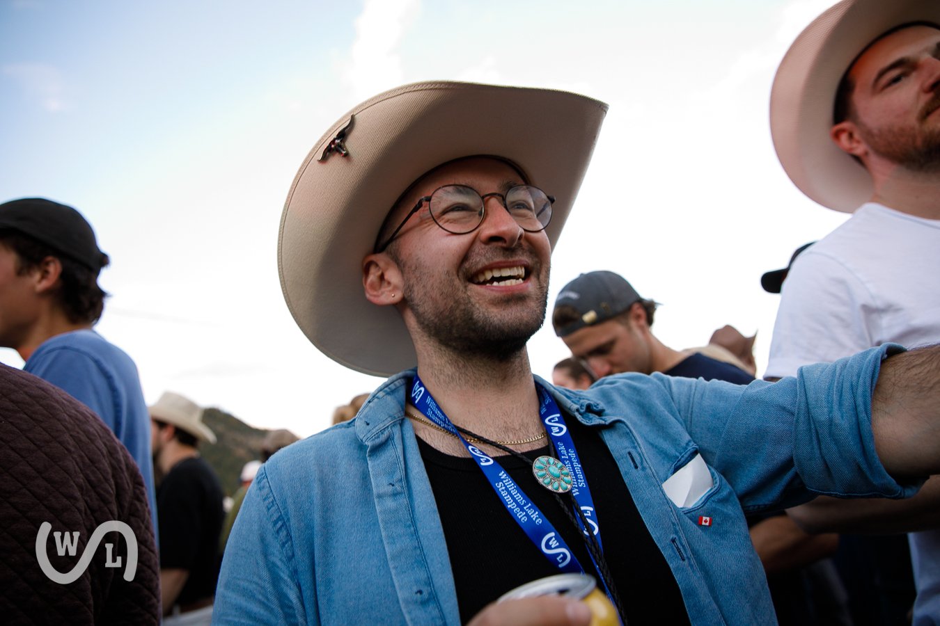 A man smiling and wearing glasses and a wide-brimmed hat at an outdoor event with other people around.