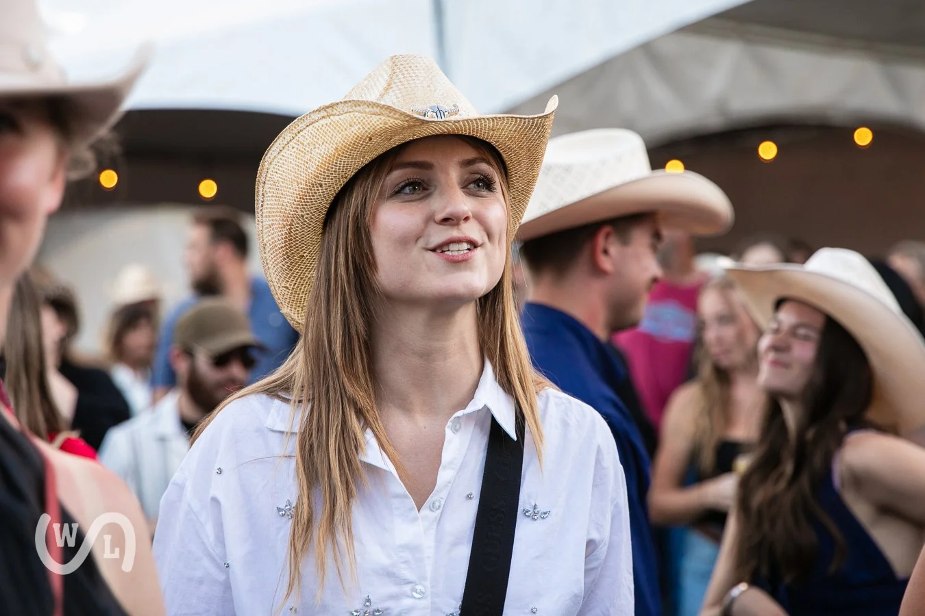 A young woman wearing a cowboy hat, white shirt, and shoulder strap at a crowded outdoor event with other people in cowboy hats and casual attire.