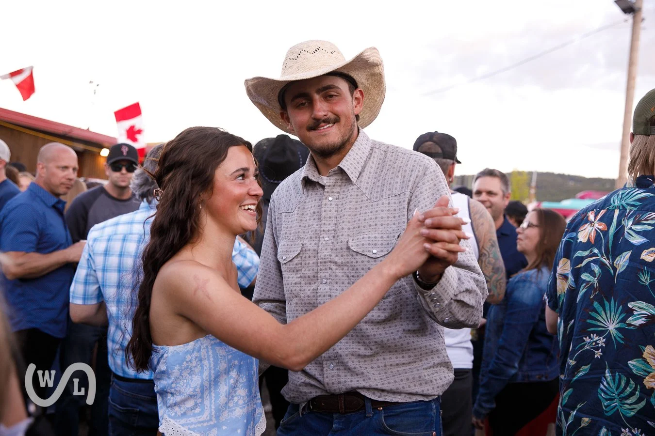 A happy couple dancing at an outdoor event with other people in the background, some holding Canadian flags.