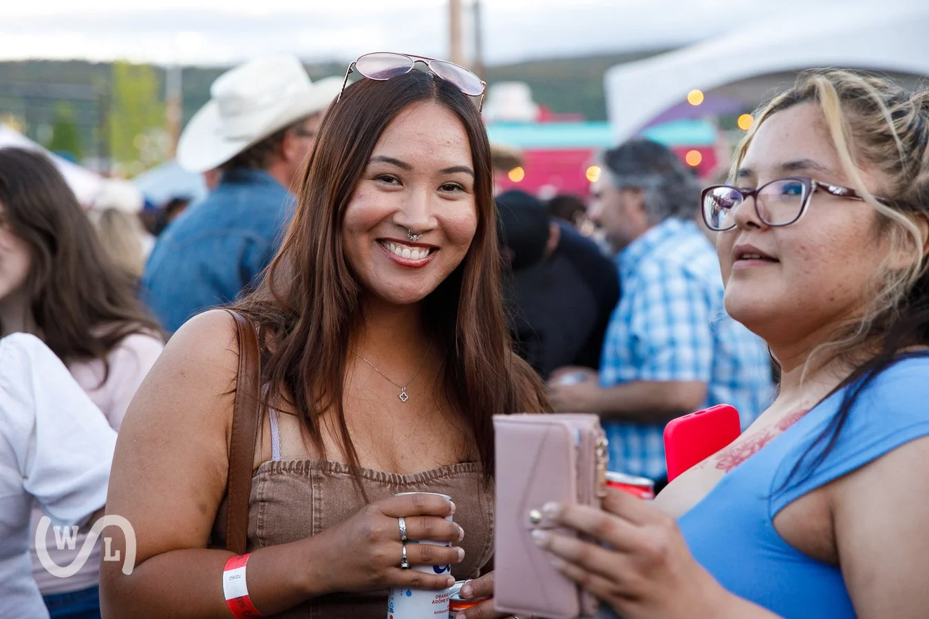 Two women at an outdoor event, smiling and talking, with a crowd and tents in the background.