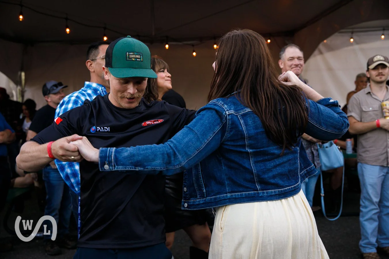 A couple dancing together at an outdoor event, surrounded by other people under string lights.