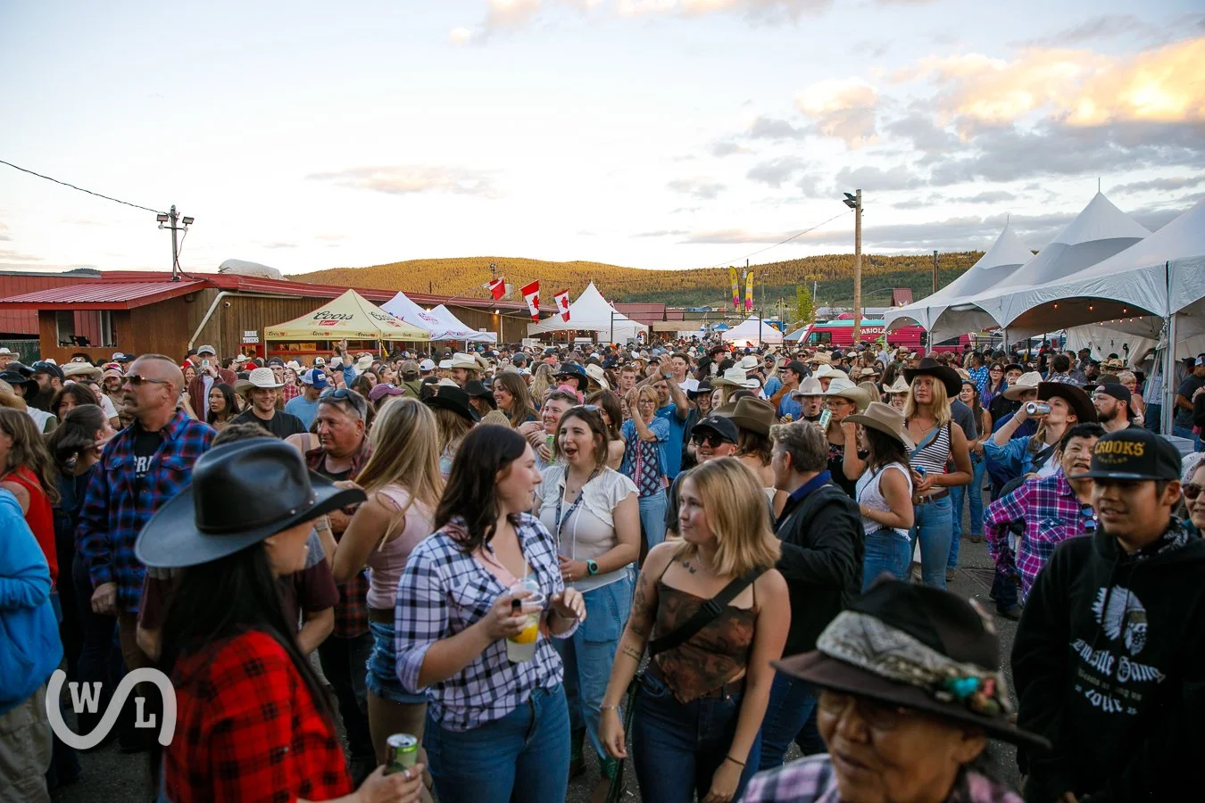 Crowd of people at an outdoor event or festival with tents and flags in the background, grassy hills under a partly cloudy sky.